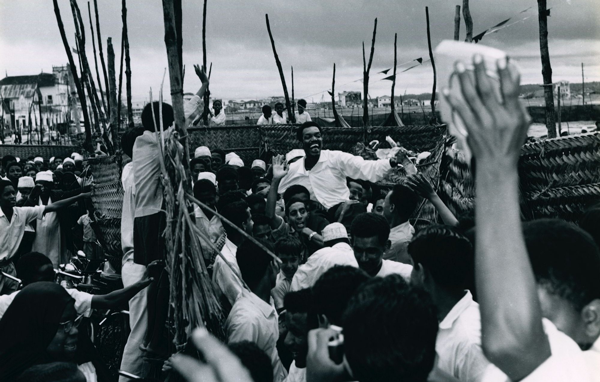 L'image montre une scène animée et dynamique, probablement lors d'un rassemblement ou d'une célébration. De nombreuses personnes se regroupent, leurs visages expriment la joie et l'excitation. On peut voir des hommes et des femmes, certains portant des vêtements traditionnels. Au centre, un homme semble chercher à atteindre un groupe, suscitant des réactions enthousiastes de la foule. Les bras de plusieurs personnes s'élèvent, clairement engagés dans l'événement. L'arrière-plan apparaît flou, avec des éléments de construction ou des arbres qui ajoutent à l'atmosphère vivante de la scène. L'ambiance générale évoque une forte connexion et une communauté vibrante.