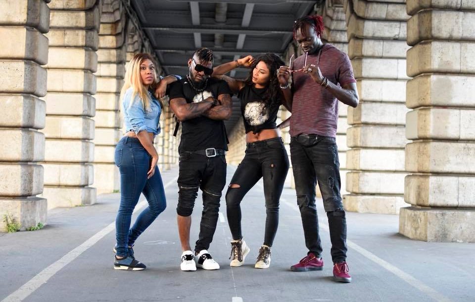 L'image présente un groupe de quatre personnes se tenant sous un pont, avec des colonnes en pierre sur les côtés. À gauche, une femme porte une chemise en denim et un jeans serré, affichant une posture confiante. À côté d'elle, un homme avec un look musclé, portant un tee-shirt noir, se distingue par ses lunettes de soleil et sa barbe. Il est entouré de deux autres femmes. L'une d'elles a les cheveux longs et noirs, vêtue d'un haut noir et d'un jeans. Elle se tient légèrement penchée, adoptant une attitude détendue. La dernière personne est un homme avec des cheveux colorés, aussi habillé de manière décontractée, tenant un appareil dans ses mains. L'environnement donne une ambiance urbaine, renforcée par le béton et les ombres créées par la structure du pont.