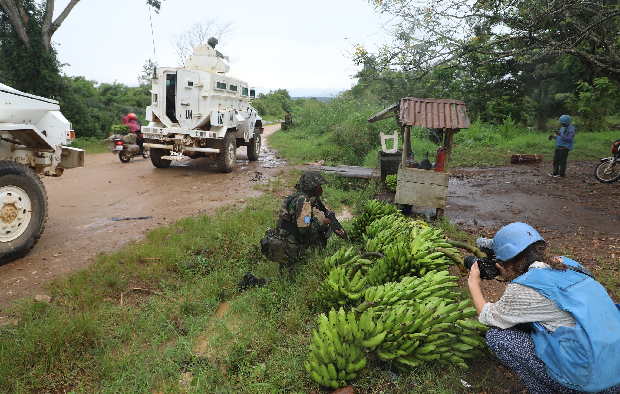 L'image dépeint une scène rurale dans un environnement naturel. Sur le côté gauche, on aperçoit un groupe de bananes bien rangées au sol, d'un vert vibrant. À proximité, un soldat en uniforme utilise une caméra pour capturer des images, se penchant légèrement vers les bananes. Sur la droite, un véhicule blindé est garé sur une route poussiéreuse, entouré d'une végétation luxuriante. Au loin, d'autres personnes, comme un homme sur une moto et une femme à pied, ajoutent une touche de vie à cette scène. Le ciel semble nuageux, suggérant un temps humide, et l'atmosphère générale évoque un mélange d'activités militaires et de vie quotidienne.