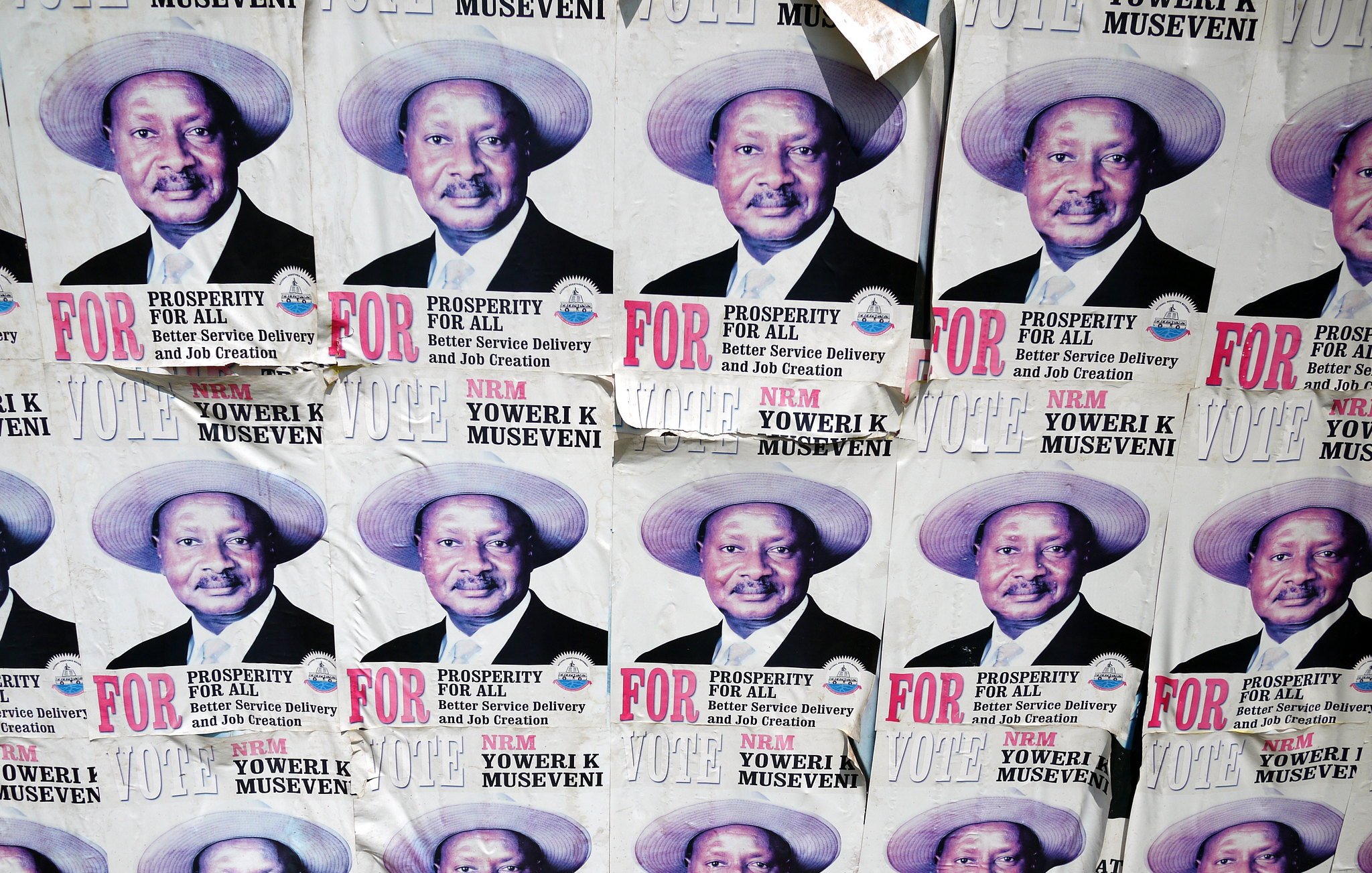 L'image montre un collage de plusieurs affiches sur un mur. Chaque affiche présente le même portrait d'un homme portant un chapeau de paille. Il a un grand sourire et affiche une expression confiante. Sous son image, on peut lire des textes en lettres majuscules qui soulignent des mots comme "PROSPERITY" (prospérité) et "FOR" (pour). Les affiches semblent faire partie d'une campagne politique, faisant appel au soutien des électeurs. Le fond est en papier décoloré et usé, indiquant que ces affiches ont été exposées pendant un certain temps.