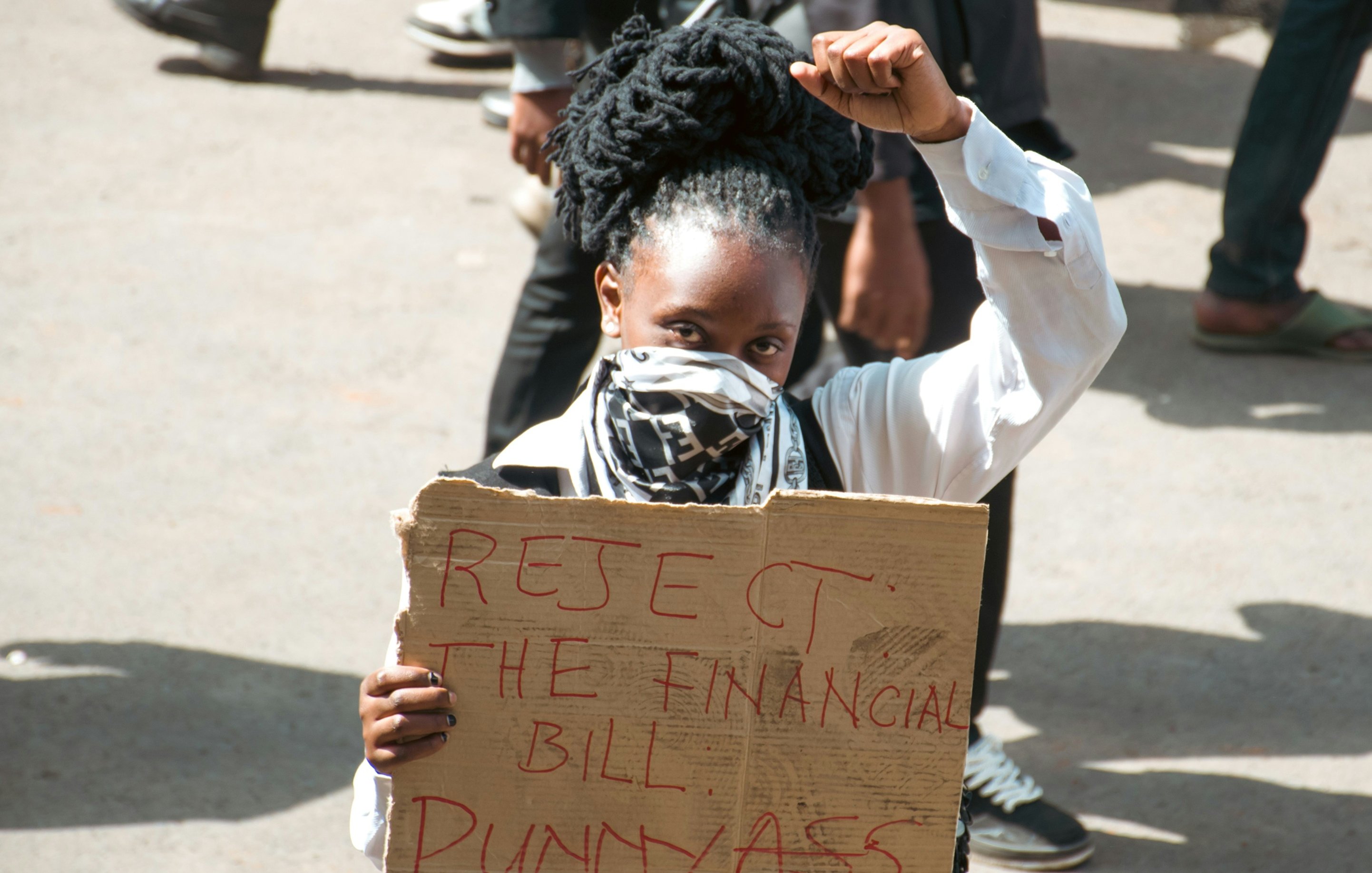 L'image montre une manifestation dans laquelle une femme se tient au premier plan. Elle a des cheveux noirs et crépus coiffés en chignon. Elle porte un masque sur le visage et un haut blanc. Dans sa main, elle élève le poing, symbole de résistance. L'autre main tient une pancarte en carton sur laquelle sont inscrits en lettres rouges les mots "REJECT THE FINANCIAL BILL PUNNYASS". En arrière-plan, on distingue d'autres personnes, soulignant le caractère collectif de la protestation. L'atmosphère est chargée d'énergie, avec un sentiment d'engagement palpable.