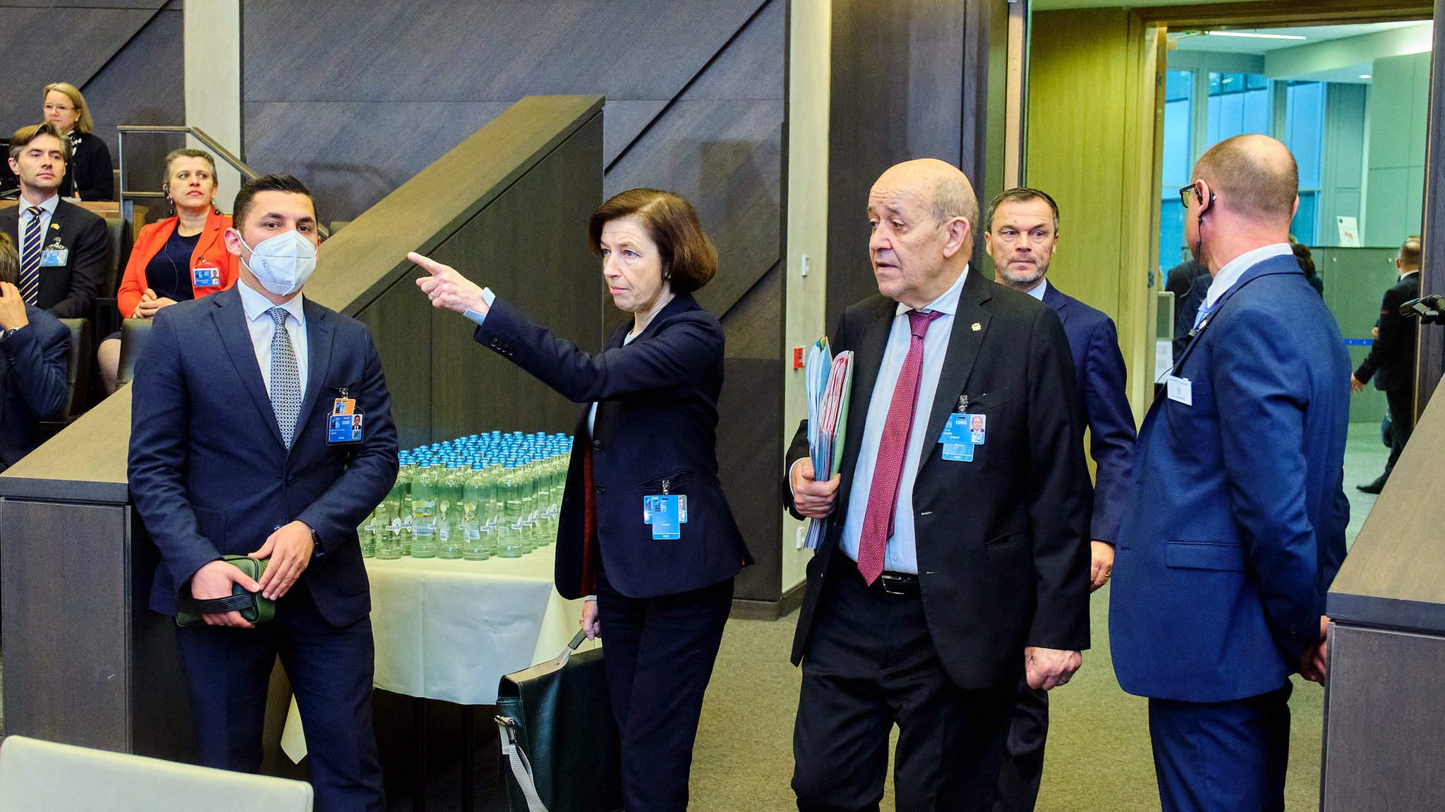 Dans cette scène, un groupe de personnes se déplace à l'intérieur d'un bâtiment moderne, probablement lors d'une réunion ou d'un événement officiel. Au premier plan, une femme, vêtue d'un costume professionnel et portant un document à la main, pointe dans une direction, semblant donner des indications. À ses côtés, un homme portant un masque de protection écoute attentivement, tandis qu'un autre homme, en costume noir, marche à ses côtés, apparemment engagé dans une conversation. En arrière-plan, d'autres participants sont visibles, suggérant une atmosphère animée et collaborative. Des bouteilles d'eau sont disposées sur une table à proximité, signalant la durée de l'événement. Le décor est contemporain, avec des murs en bois et des éléments de design épuré.