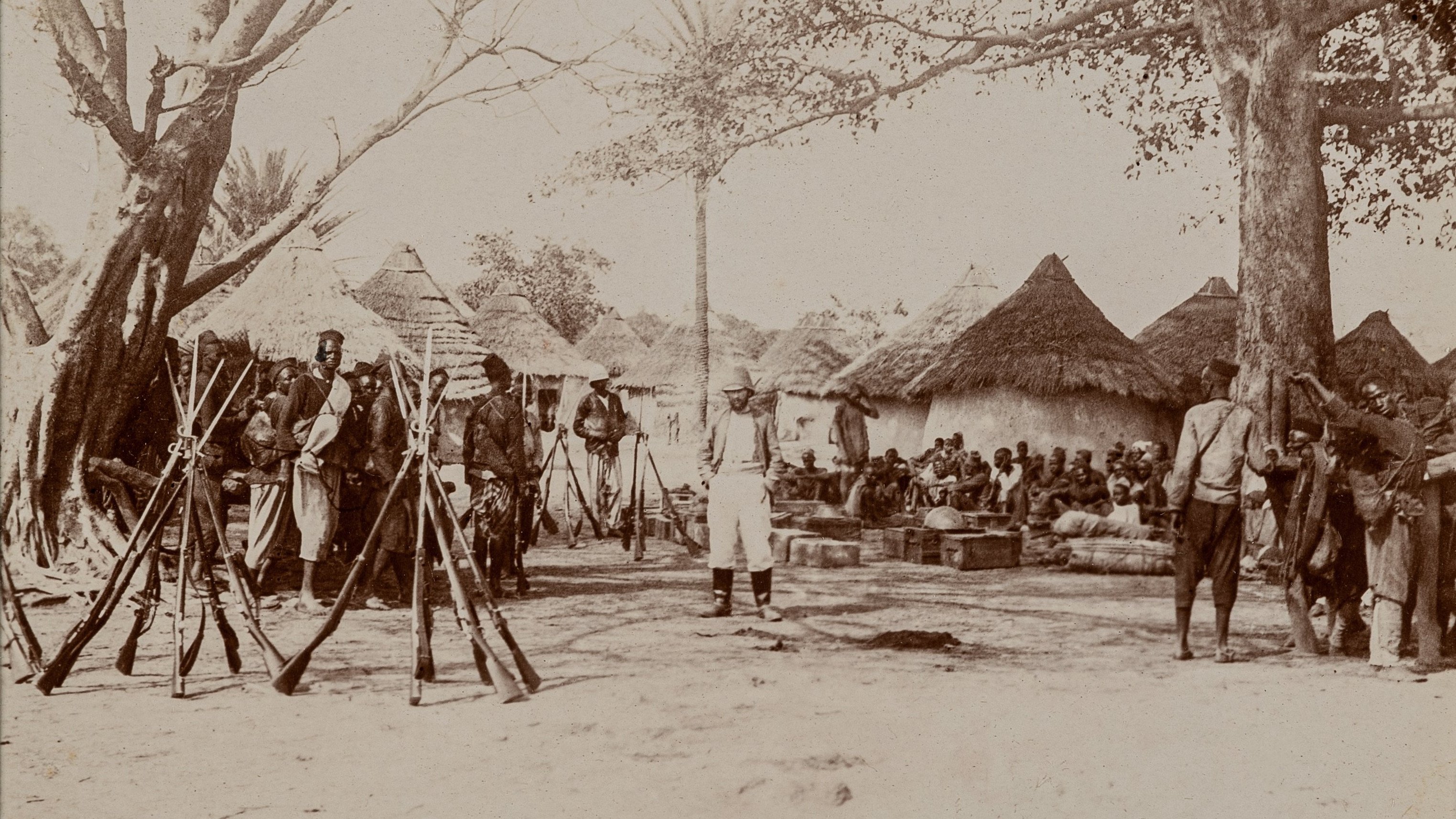 L'image représente une scène historique à l'extérieur d'un village. Au premier plan, plusieurs fusils sont posés verticalement sur le sol, formant une sorte de cadre. Des hommes, en majorité vêtus de vêtements traditionnels, sont rassemblés autour d'une zone centrale. Certains semblent discuter tandis que d'autres restent attentifs, leur posture indiquant un moment de rassemblement communautaire ou de célébration. En arrière-plan, on aperçoit des huttes de forme circulaire, couvertes de toits en paille, typiques des constructions de certaines régions rurales. Plusieurs arbres offrent de l'ombre, ajoutant à l'ambiance naturelle de l'endroit. Le sol est poussiéreux, indiquant le climat aride de la région. Les expressions des visages reflètent un mélange d'intérêt et de sérieux, suggérant une rencontre importante au sein de la communauté.