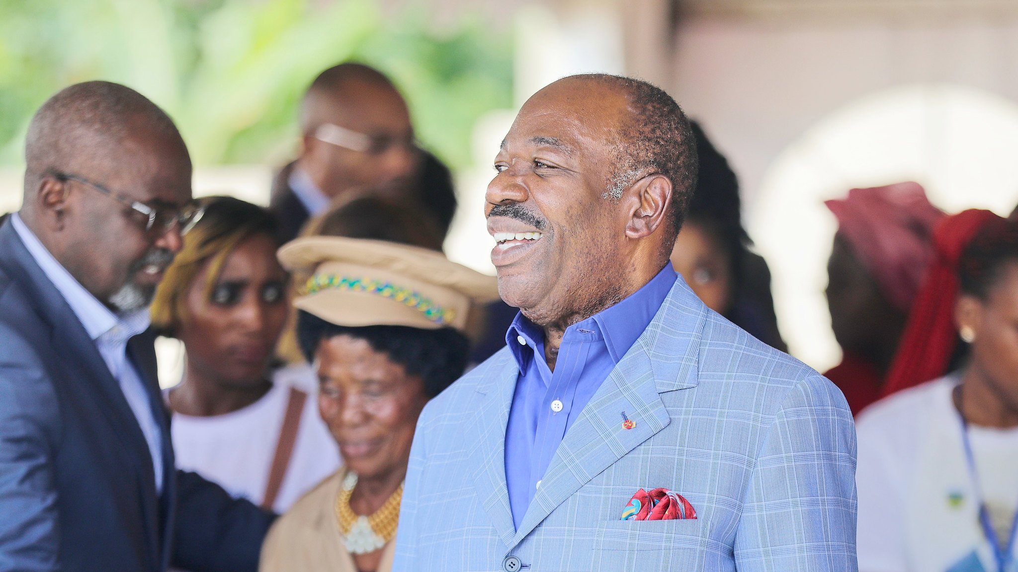L'image montre un groupe de personnes en train d'interagir dans un cadre convivial. Au centre, un homme, portant un costume bleu clair et une chemise bleue, sourit chaleureusement, affichant une attitude amicale et accessible. On note également des personnes en arrière-plan, certaines portant des vêtements colorés, qui semblent engagées dans une conversation ou une activité sociale. L'ambiance globale de l'image est joyeuse et accueillante, suggérant un moment de célébration ou de rencontre.