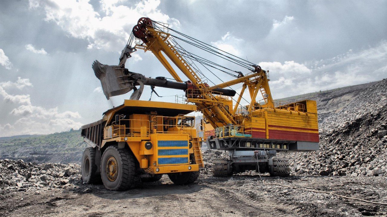 L'image présente un site d'extraction minière. Au premier plan, un gros camion de transport de couleur jaune, avec des roues massives, est chargé de matériaux. À côté, une grande pelle mécanique jaune et rouge, dotée d'un bras articulé, a relevé une charge de terre ou de roches. Le paysage environnant est accidenté, avec des tas de gravier et des pentes abruptes. Le ciel est partiellement nuageux, laissant passer des rayons de lumière qui illuminent la scène, créant une ambiance industrielle dynamique. Des bruits de machines et d'activités minières peuvent être imaginés, rendant l'environnement à la fois impressionnant et robuste.