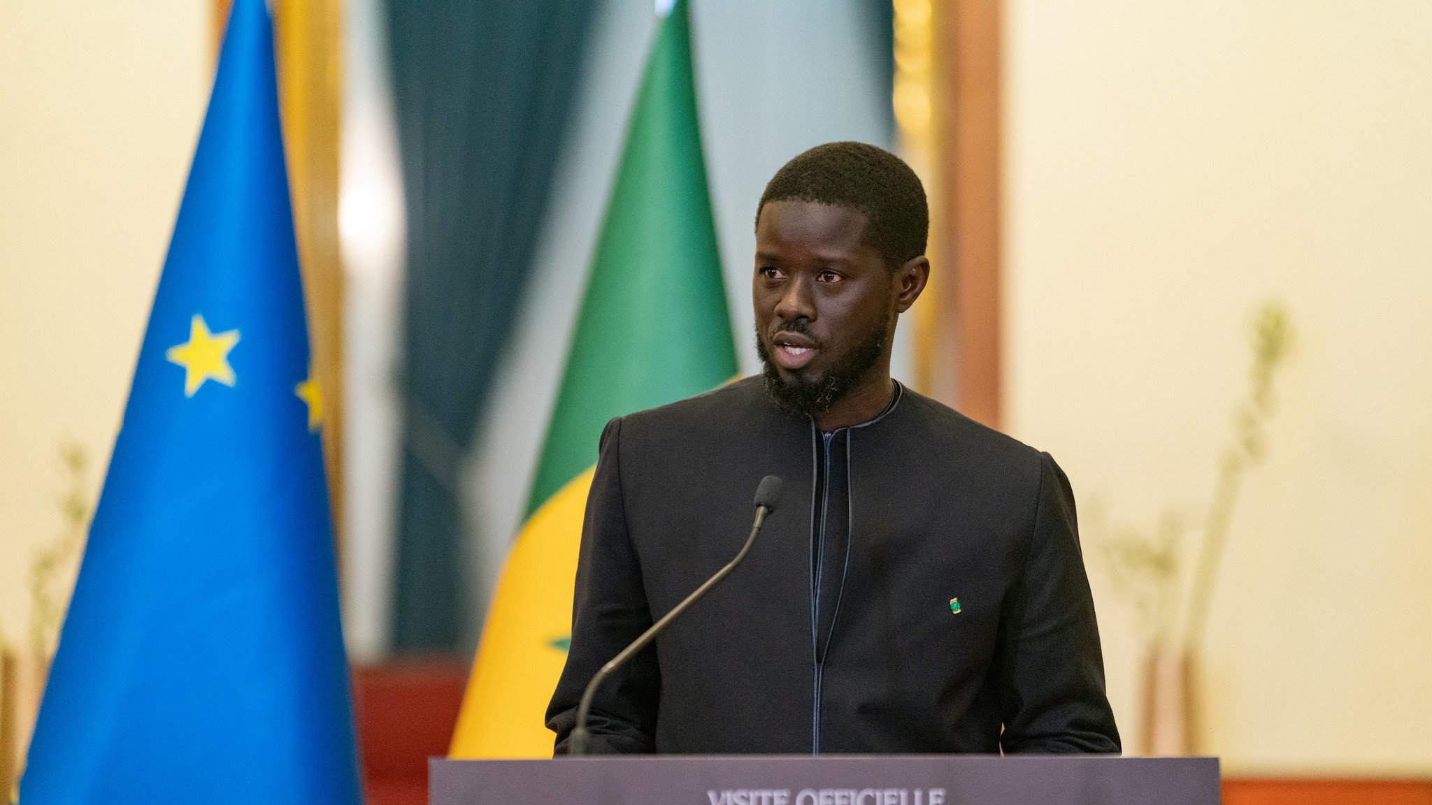 Sur cette image, un homme est en train de prendre la parole lors d'une conférence. Il est vêtu d'une tenue traditionnelle noire et se tient devant un podium portant une plaque avec des inscriptions officielles. En arrière-plan, on peut voir deux drapeaux : l'un affiche les étoiles de l'Union européenne et l'autre représente le Sénégal. L'homme semble sérieux et engagé, s'adressant à un public. La scène dégage une atmosphère formelle et diplomatique.