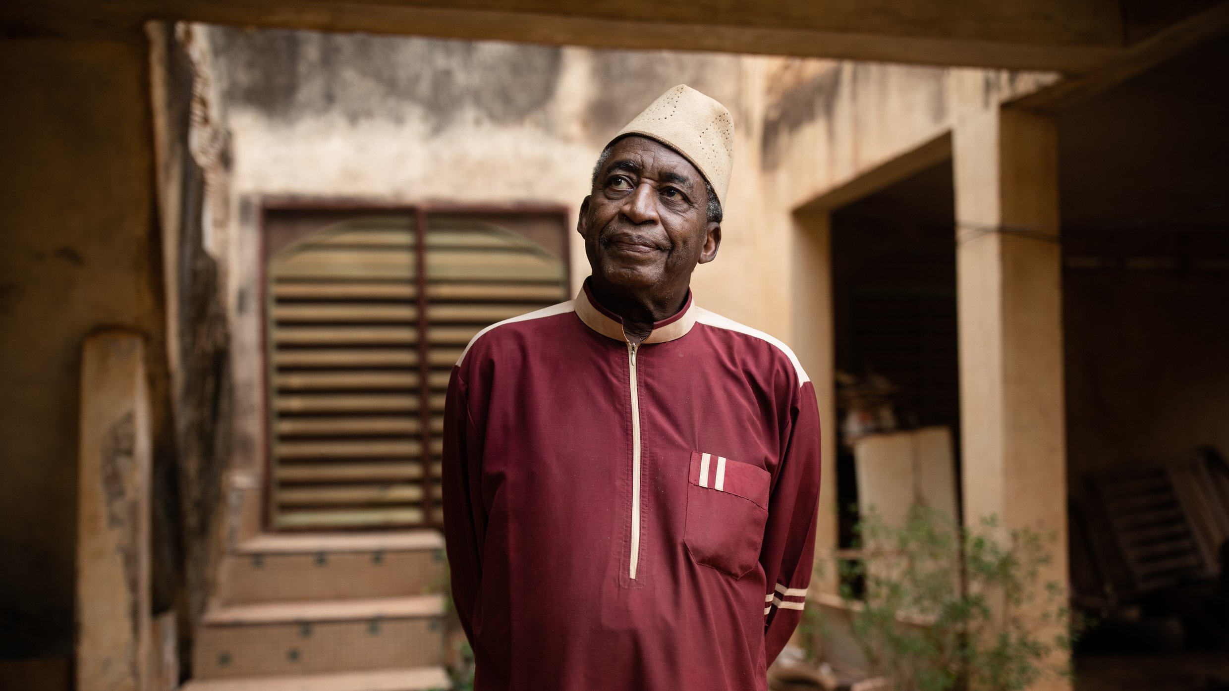L'image montre un homme âgé portant une tenue traditionnelle d'une couleur rouge foncé. Sa chemise longue est agrémentée de détails blancs. Il se tient debout, les bras le long du corps, et regarde légèrement vers le haut. L'arrière-plan présente un bâtiment avec des murs usés et une porte en bois, ainsi que quelques plantes verdoyantes qui ajoutent une touche de nature à la scène. L'homme dégage une expression calme et réfléchie, créant une atmosphère de sagesse et de sérénité.