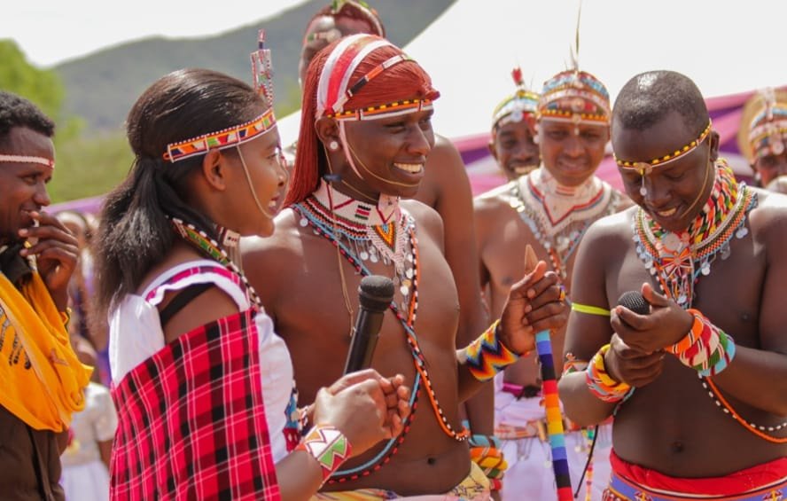 Dans cette image, un groupe de personnes participe à une cérémonie culturelle vibrante. Au premier plan, une femme, vêtue d'une robe traditionnelle colorée avec des motifs, tient un micro et s'adresse à un homme qui se tient à côté d'elle. Cet homme porte un pagne traditionnel et est orné de nombreux accessoires en perles. Ils sont tous deux entourés de plusieurs autres hommes, également en tenue traditionnelle, qui sourient et semblent s'engager dans une conversation joyeuse. Les vêtements sont décorés de motifs colorés et de bijoux en perles, ce qui évoque une atmosphère festive. L'arrière-plan montre des collines verdoyantes, et l'ambiance générale dégage un sens de communauté et de célébration de la culture. L'image respire l'énergie et la convivialité, illustrant des rituels ou des danses traditionnelles.