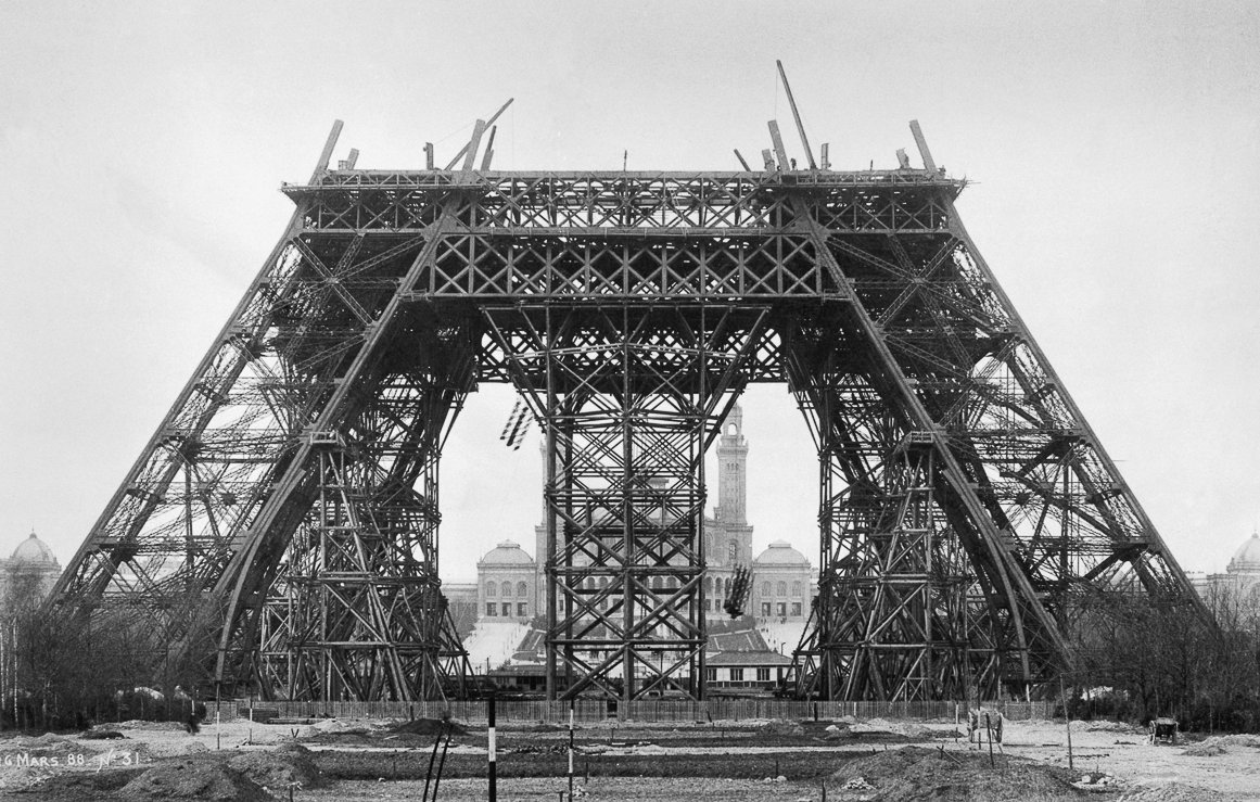 Cette image montre une phase de construction de la célèbre Tour Eiffel à Paris. On peut voir une structure métallique imposante, caractérisée par des poutres en fer entrecroisées. La tour, encore incomplète, présente une base large et stable avec des arcades qui s'élèvent vers le ciel. À l'arrière-plan, on distingue un bâtiment, peut-être une mairie, qui ajoute un contexte urbain à la scène. L'ambiance globale de l'image est monochrome, ce qui renforce l'aspect historique et donne un aperçu des débuts de cette icône de l'architecture. Le sol est encore en chantier, avec des débris et des outils, ce qui rappelle le processus de construction en cours.