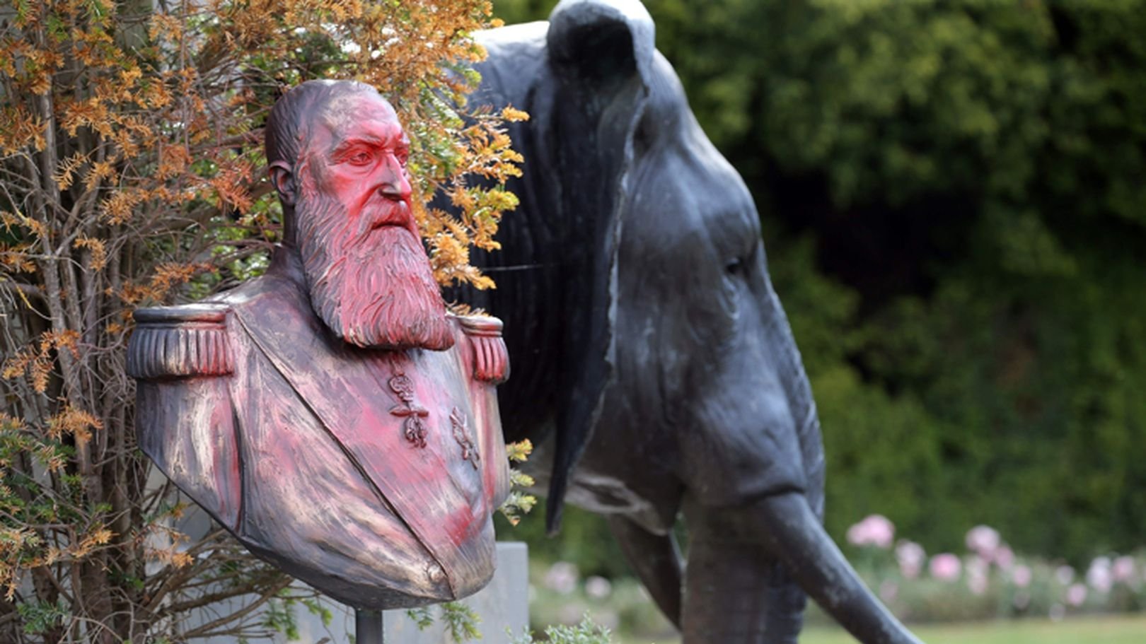 L'image présente un buste en bronze d'un homme barbu, aux cheveux longs et portant des uniformes militaires, posé sur un socle. Ce buste est entouré de végétation, ce qui lui donne un aspect naturel. En arrière-plan, on aperçoit la silhouette d'un éléphant en métal, affichant des détails réalistes comme ses grandes oreilles et sa trompe. Le contraste entre le buste humain et l'éléphant, ainsi que la verdure environnante, crée une ambiance paisible et mémorable dans un jardin.