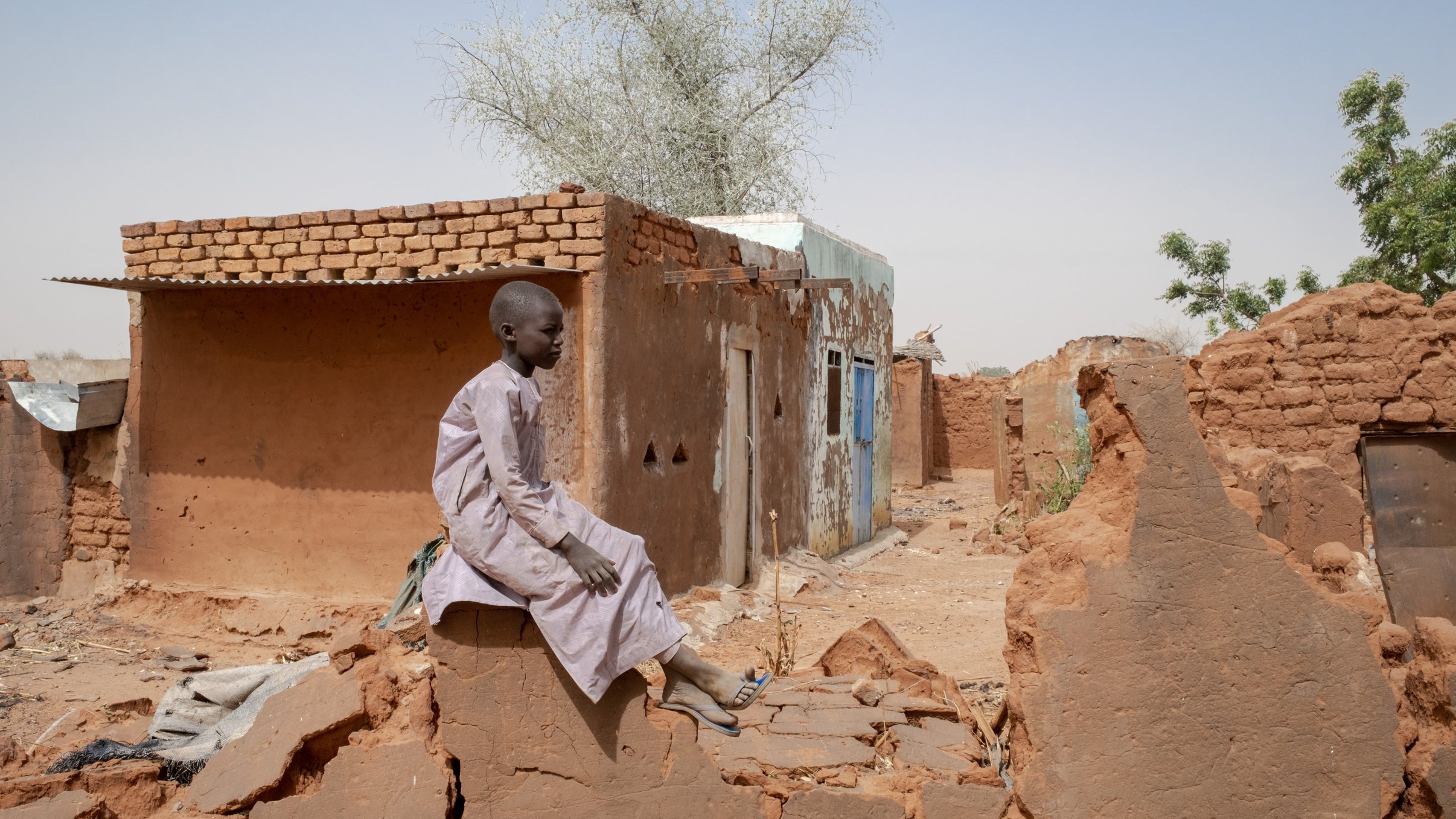 L'image montre un jeune garçon assis sur un mur de terre, au milieu d'un paysage rural aride. Il porte une longue tunique claire et semble pensif. En arrière-plan, on voit des maisons en terre battue, avec des murs qui sont fissurés ou partiellement détruits. L'environnement est sec, avec quelques arbres isolés. Le ciel est dégagé, d'un bleu clair. L'atmosphère générale transmet une sensation de solitude et de résilience dans un milieu rural difficile.