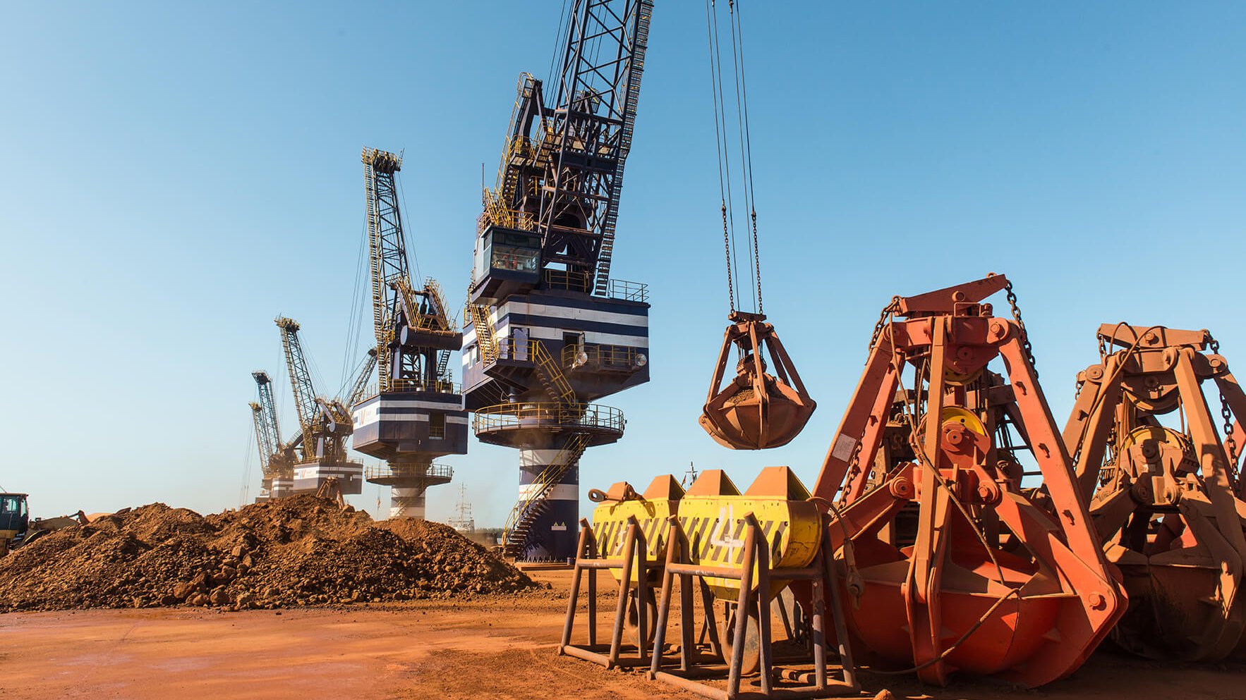 Dans cette image, nous sommes dans un port industriel où plusieurs grues sont visibles. Ces grandes machines sont utilisées pour charger et décharger des matériaux. À l'avant, il y a des godets et des bennes orange, qui sont des équipements de levage, reposant sur le sol. En arrière-plan, plusieurs grues se dressent contre un ciel bleu clair, leurs bras et leurs crochets descendus vers les tas de terre brune. L'atmosphère est typiquement industrielle, avec un sol en terre battue et une ambiance de travail intense. Des sons de machines en fonctionnement et le bruit des cargaisons en mouvement pourraient être présents dans cet environnement.