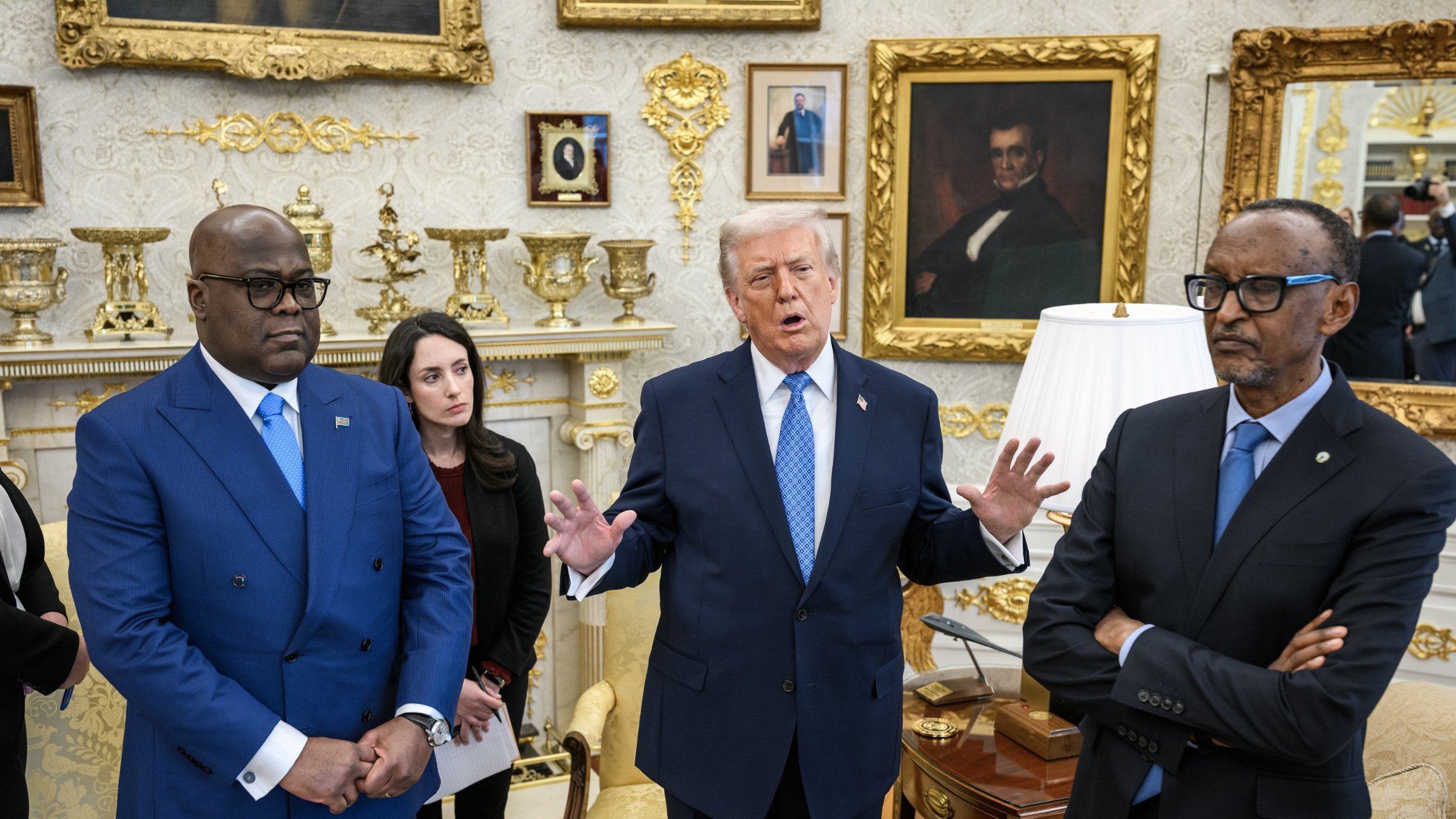 Dans cette image, nous sommes dans le Bureau Ovale, un espace formel et décoré richement. Au centre, un homme, probablement une figure politique, gesticule en s'exprimant avec animation. À sa gauche, un homme en costume bleu écoute attentivement, tandis qu'à droite, un autre homme, également en costume sombre, semble plus réservé, les bras croisés. Une femme en arrière-plan prend des notes, portant un vêtement noir. L'ambiance de la scène montre des échanges significatifs, probablement liés à des discussions politiques ou diplomatiques. Les murs sont ornés de cadres dorés, avec des portraits et des éléments décoratifs qui ajoutent à l'élégance de la pièce.