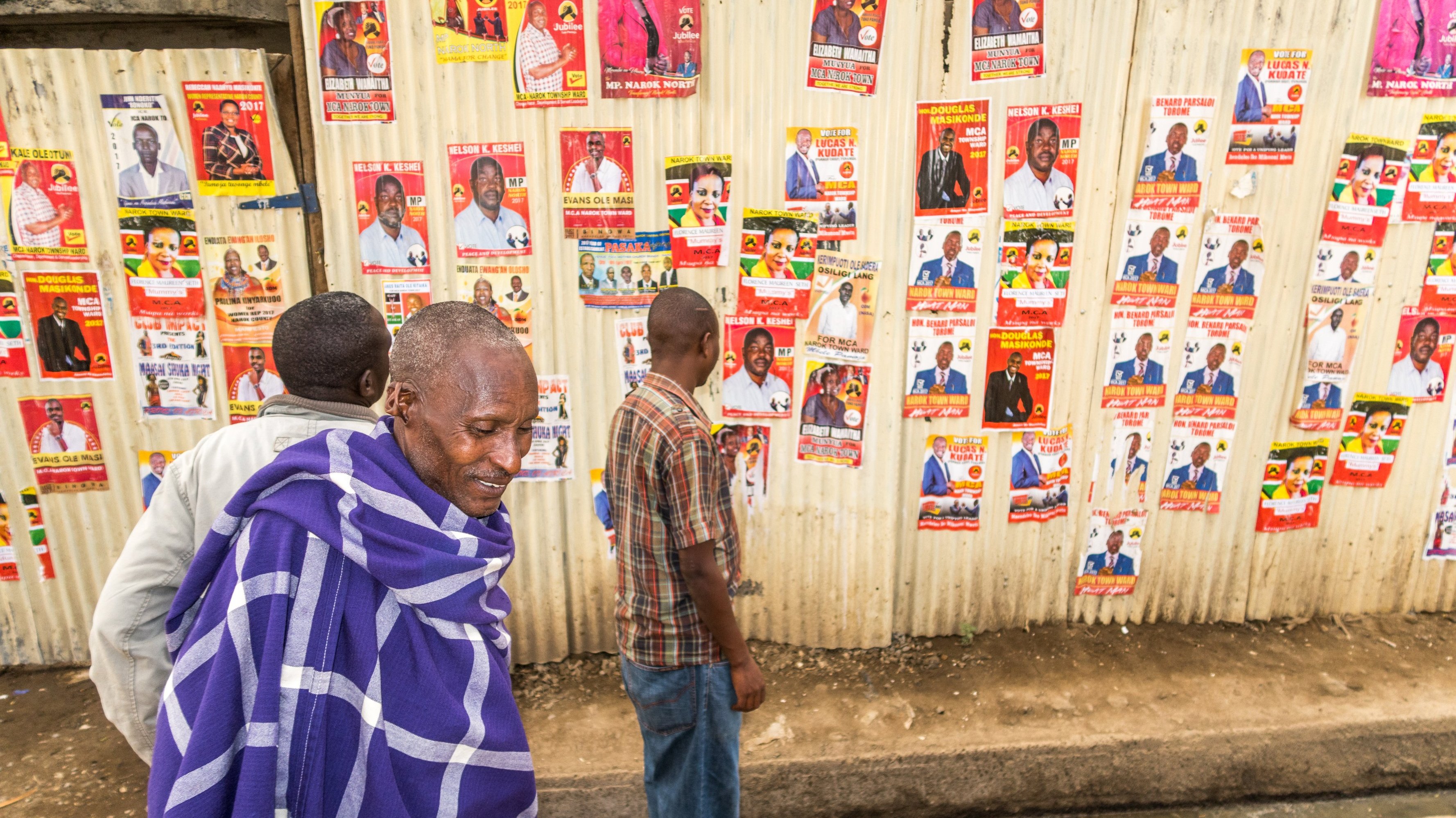 L'image montre un groupe de personnes marchant le long d'un mur recouvert de nombreuses affiches. Ce mur en tôle est tapissé de posters colorés présentant des candidats politiques, avec des visages souriants et des informations écrites. Au premier plan, un homme est vêtu d'un châle traditionnel bleu et blanc, semblant contempler les affiches. À ses côtés, d'autres personnes se déplacent lentement, examinant les posters tandis que le sol est pavé, dans un environnement urbain. L'atmosphère semble à la fois animée et réfléchie, avec un mélange de culture locale et d'engagement politique.