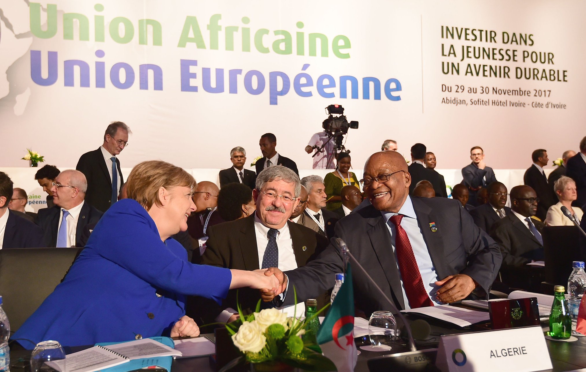 L'image montre la scène d'une conférence internationale où des dirigeants participent à un sommet. Au premier plan, on aperçoit un groupe de personnes, parmi lesquelles une femme, identifiée comme une figure politique importante, est en train de serrer la main d'un homme. Cette interaction symbolise le dialogue et la collaboration entre les nations. À l'arrière-plan, on distingue des bannières et des affiches portant le titre "Union Africaine, Union Européenne" ainsi que le slogan "Investir dans la jeunesse pour un avenir durable". La salle semble remplie de délégués, en costard et concentrés sur les discussions, ce qui donne une atmosphère formelle et engagée. Des éléments comme une table avec des drapeaux de différents pays ajoutent au cadre diplomatique de l'événement.