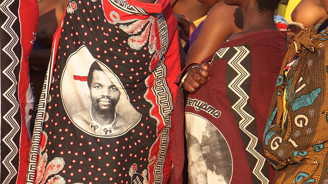 Dans cette image, on voit un groupe de femmes habillées de façon traditionnelle, revêtues de vêtements colorés et ornés de motifs riches. Certaines portent des châles qui affichent des portraits d'un homme, probablement une figure importante de leur culture. Les femmes se tiennent proches les unes des autres, exprimant un esprit de solidarité. L'atmosphère est vibrante, suggérant une célébration ou un événement culturel, avec des couleurs vives qui évoquent une culture riche et dynamique. Les sons environnants pourraient comprendre des musiques folkloriques et des rires, ajoutant à l'ambiance festive.