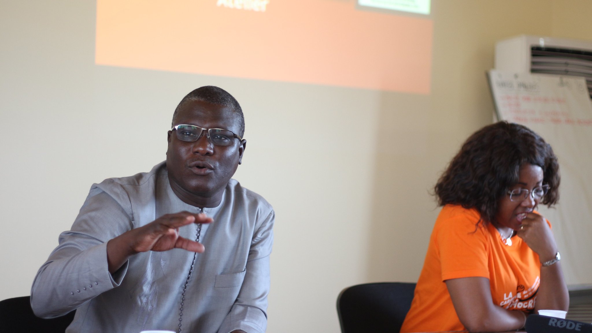 L'image montre un atelier où deux personnes sont assises à une table. À gauche, un homme, portant des lunettes et une tenue traditionnelle grise, gesticule avec une main tout en parlant. Son expression est engageante et dynamique, suggérant qu'il s'adresse à un groupe. À droite, une femme, habillée d'un t-shirt orange, écoute attentivement. Son visage montre une attitude attentive et intéressée. En arrière-plan, un écran est visible avec des textes, mais il est flou. L'atmosphère semble interactive et collaborative, indiquant un échange d'idées lors de l'atelier.