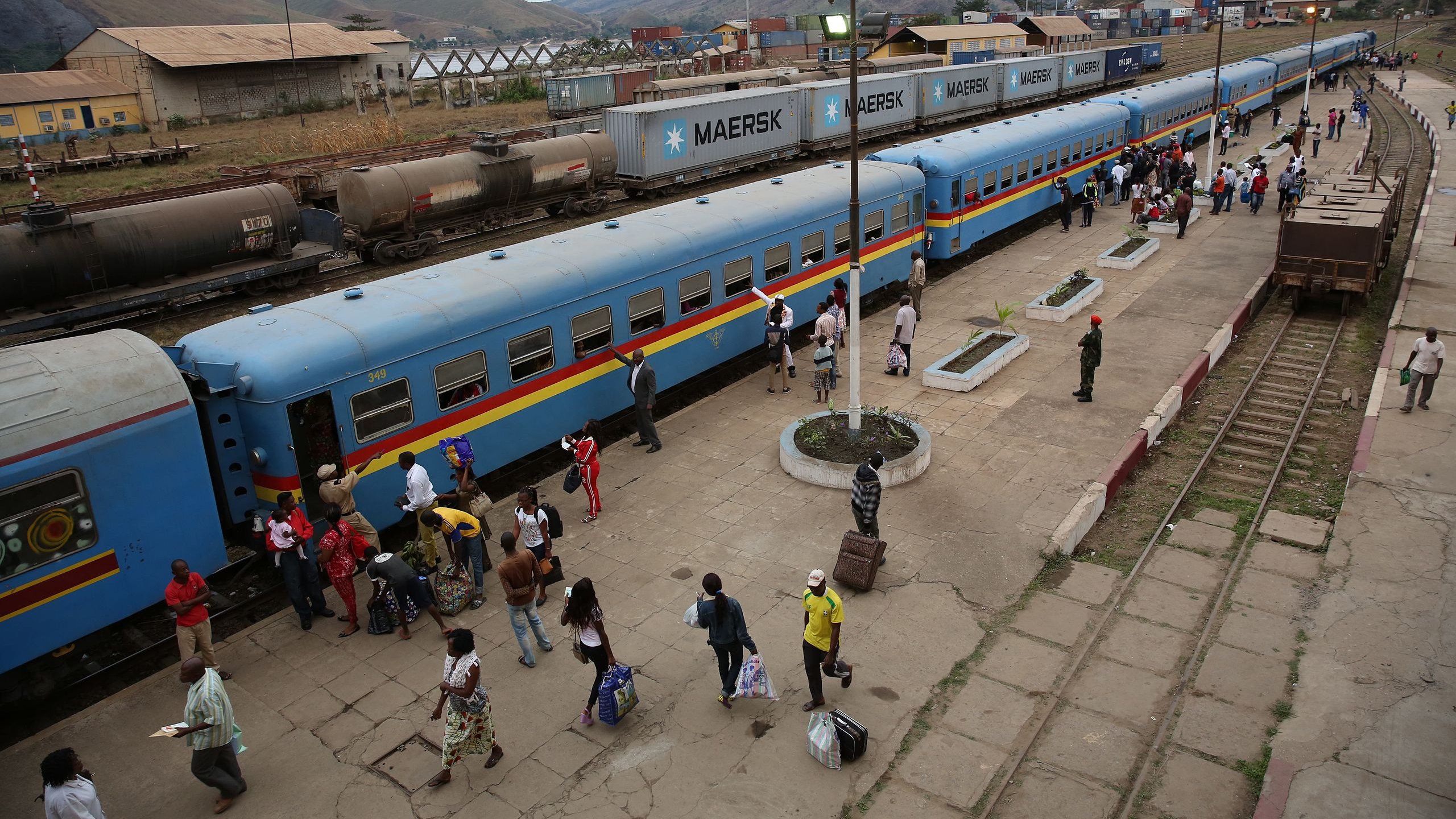 L'image montre une gare animée. De longs trains de marchandises et de passagers sont alignés sur les quais. Les wagons sont d'un bleu vif avec des bandes colorées. Des voyageurs descendent et montent à bord, portant des sacs et des effets personnels. On peut entendre des conversations, des annonces de train, et le bruit des roues sur les rails. En arrière-plan, on aperçoit des collines qui entourent la gare. L'atmosphère est vive et dynamique, avec un mélange de mouvements et d'interactions entre les passagers.