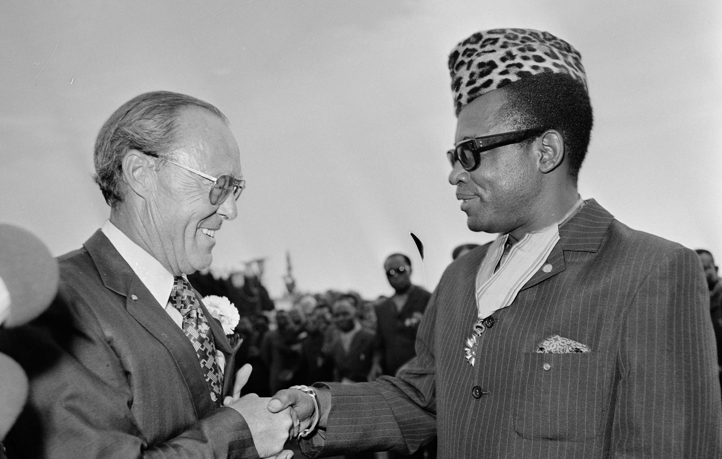 L'image montre une scène de rencontre entre deux hommes souriants, se serrant la main dans un cadre formel et convivial. Le premier homme porte un costume sombre avec une chemise à motifs fleuris et des lunettes, tandis que le second, qui semble être une figure importante, arbore un élégant costume rayé et un chapeau à motifs distinctifs, évoquant une riche culture. En arrière-plan, un groupe de personnes observe la scène, ajoutant à l'impression d'un événement important. L'atmosphère est chaleureuse et amicale, suggérant une rencontre diplomatique ou un événement marquant.
