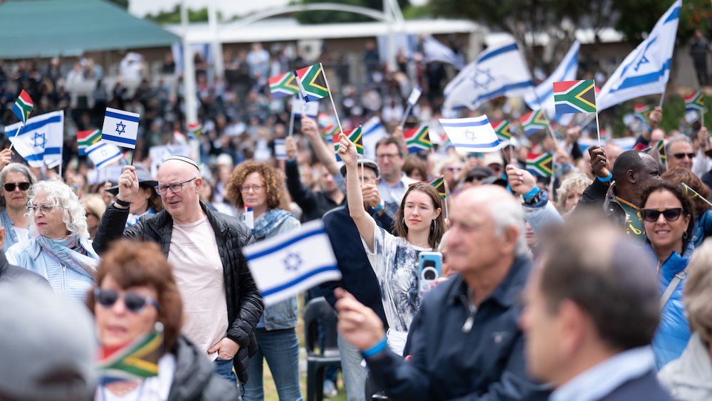 L'image montre une grande foule de personnes rassemblées dans un espace extérieur. Les gens tiennent des drapeaux, principalement des drapeaux israéliens, qui sont blancs et bleus avec l'Étoile de David, ainsi que des drapeaux sud-africains, qui présentent des couleurs vives. L'atmosphère est festive et engagée, avec des visages souriants et des expressions de joie. En arrière-plan, on peut apercevoir des assises et des personnes rassemblées autour d'un événement, suggérant une célébration ou une manifestation significative. Les vêtements des participants sont variés, allant de tenues décontractées à plus habillées, illustrant la diversité de la foule.