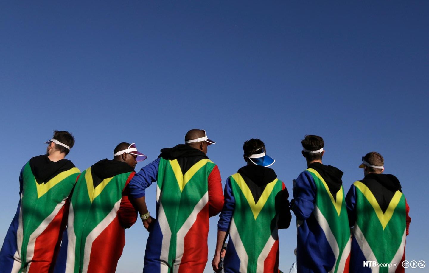 L'image montre un groupe de personnes de dos, habillées dans des capes aux couleurs du drapeau sud-africain, dominant les tons vert, noir, jaune et rouge. Leurs silhouettes sont nettement visibles contre un ciel bleu clair. Les individus semblent détendus et peuvent être en train de profiter d'un événement ou d'une célébration. Certains portent des visières sur la tête, ajoutant une touche festive à leur tenue. L'ensemble dégage une ambiance dynamique et communautaire, soulignant la fierté et l'identité culturelle.