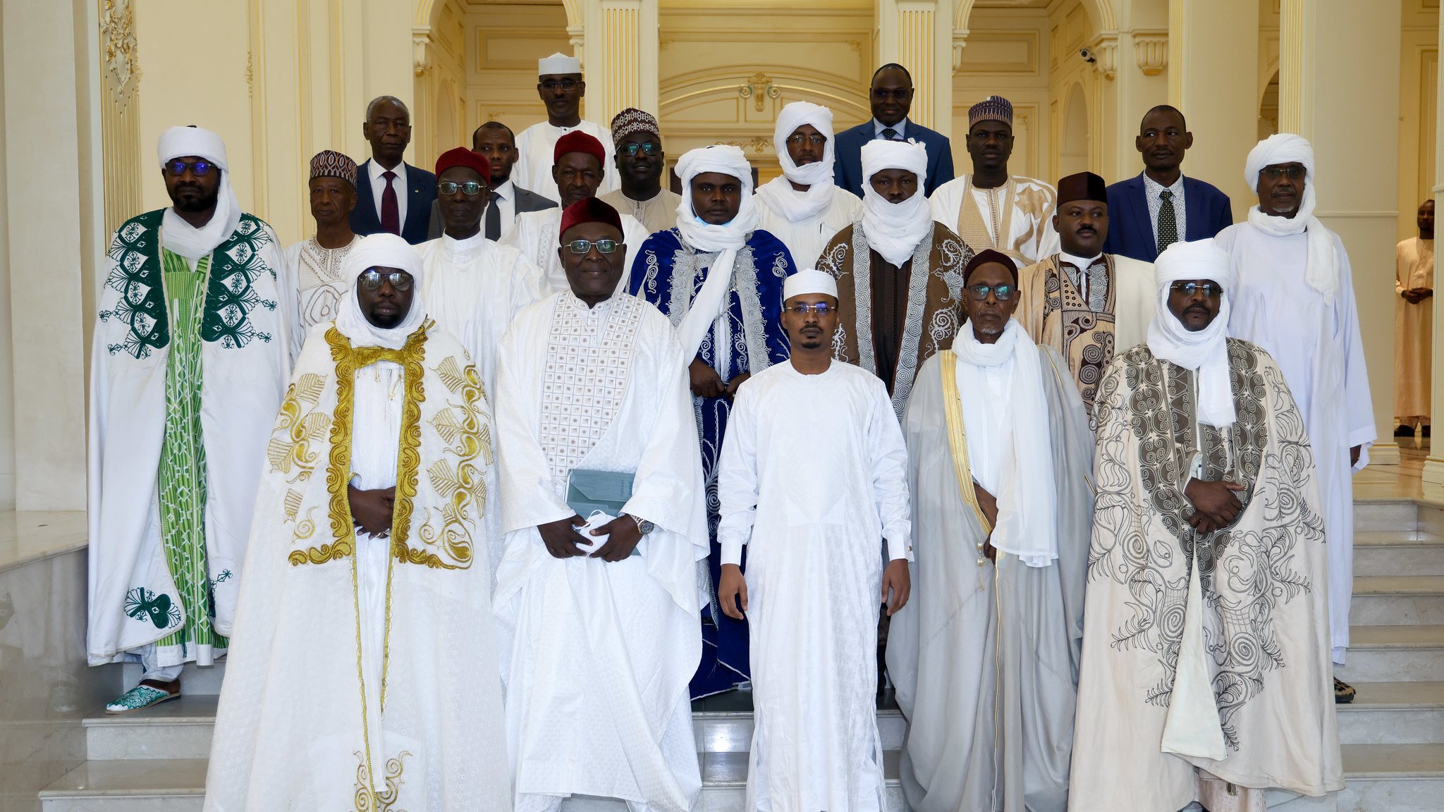 L'image montre un groupe de personnes habillées en vêtements traditionnels, rassemblées dans un espace élégant. Ils sont principalement vêtus de robes blanches, avec des éléments colorés comme des capes vertes et bleues. Les hommes portent des turbans et des chapeaux, ajoutant à la richesse de leurs tenues. L'arrière-plan est un intérieur lumineux et spacieux, avec des colonnes et des décorations architecturales. Le groupe semble coordonné et solennel, probablement pour une occasion spéciale.