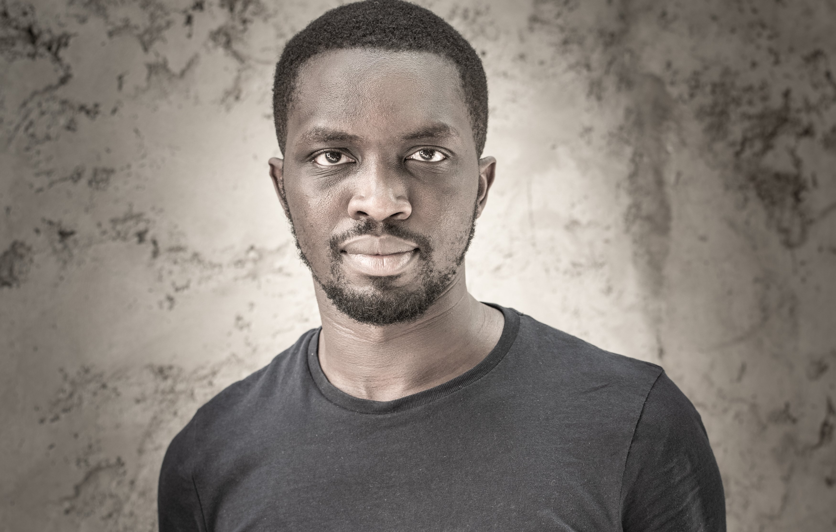 Cette image présente un homme debout, portant un t-shirt noir. Il a la peau foncée et un style de coiffure court. Son expression est sérieuse, et il regarde directement vers l'objectif avec une intensité qui attire l'attention. Le fond de l'image est texturé et neutre, mettant en valeur son visage et son corps. La lumière éclaircit certaines zones, ajoutant de la profondeur et du contraste à l'ensemble.
