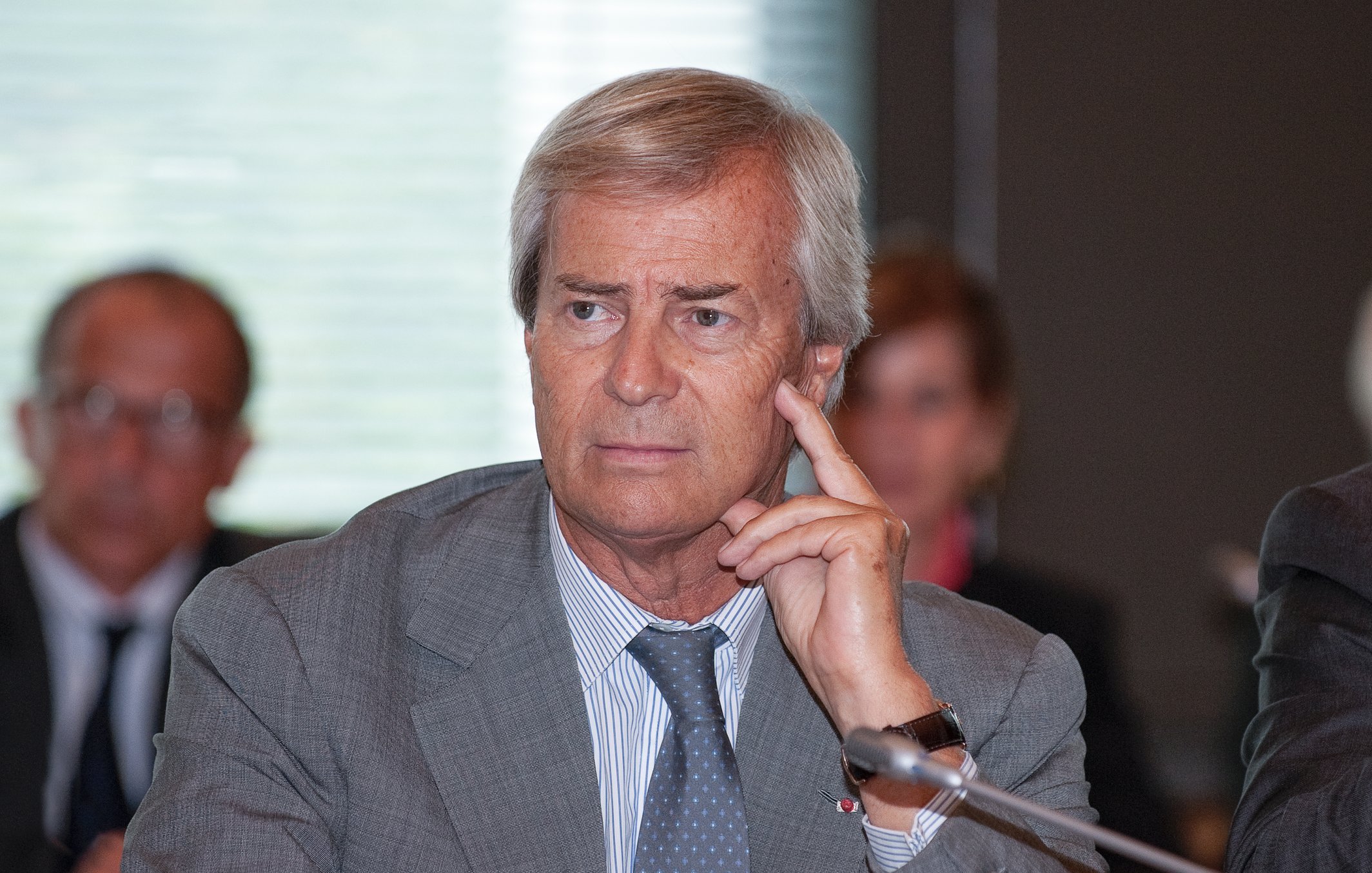 L'image montre un homme d'âge moyen, assis à une table de conférence. Il a les cheveux gris et porte un costume gris. Son expression est pensive, et il touche son visage avec un doigt en réfléchissant. En arrière-plan, on peut distinguer d'autres personnes assises, mais leurs visages sont flous. L'ambiance semble professionnelle, suggérant une réunion ou une discussion importante. La lumière est douce, créant une atmosphère de concentration.