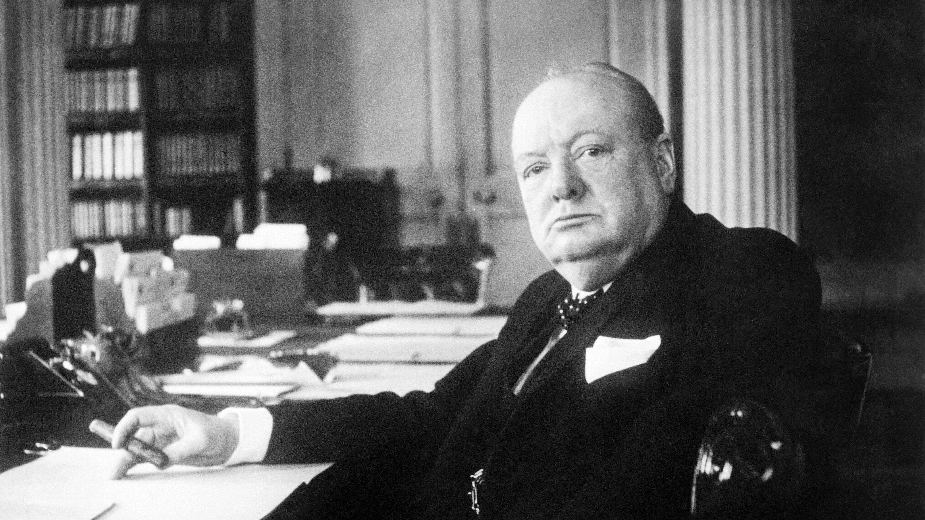 L'image présente un homme assis à un bureau. Il a une expression sérieuse et regarde droit devant lui. Son visage est rond et bien en chair, avec des traits marqués. Il est vêtu d'un costume sombre, avec une chemise claire et une cravate. Dans sa main, il tient un stylo, et devant lui, des feuilles de papier sont disposées sur le bureau. En arrière-plan, on distingue des étagères remplies de livres. L'atmosphère de l'image suggère une ambiance de travail studieuse et normalement formelle.