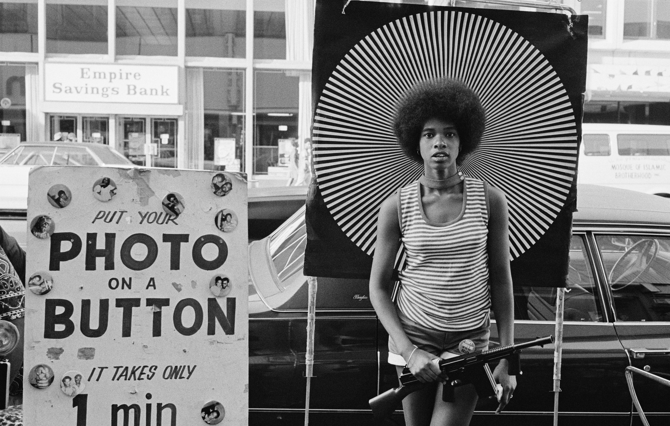 L'image présente une scène urbaine en noir et blanc. Au premier plan, une jeune femme avec une coiffure afro se tient de manière confiante, tenant une arme à feu. Elle porte un haut à rayures et se tient devant un arrière-plan décoré de motifs en spirale noirs et blancs, ce qui attire l'œil et crée un contraste frappant. À sa gauche, un panneau indique "PUT YOUR PHOTO ON A BUTTON" avec du texte dans une police simple, ajoutant un contexte de marché ou d'événement communautaire. En arrière-plan, on peut apercevoir des bâtiments et des véhicules, suggérant une atmosphère animée de la ville. L'image évoque des thèmes de pouvoir, de culture et de contestation.