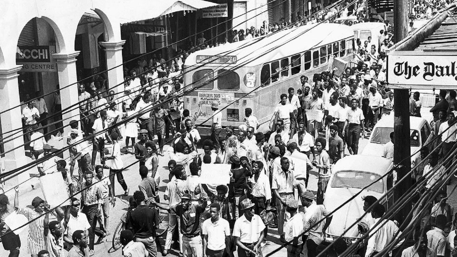 L'image que vous avez devant vous représente une scène urbaine animée dans les années 1960. On peut voir une foule dense de personnes marchant dans une rue. Les gens portent des vêtements typiques de l'époque, et il semble qu'ils se déplacent avec un but précis. À gauche, plusieurs commerces sont visibles, avec des enseignes en lettres visibles. Un ancien bus à l'allure caractéristique est garé, ajoutant une touche nostalgique à la scène. On aperçoit également quelques voitures stationnées et d'autres qui circulent dans la rue. Les fils électriques sont visibles en haut de l'image, accentuant l'impression d'une ville vivante et dynamique. L'atmosphère générale évoque une période de mouvement socioculturel important, avec des échanges et une vitalité palpable dans l'air.