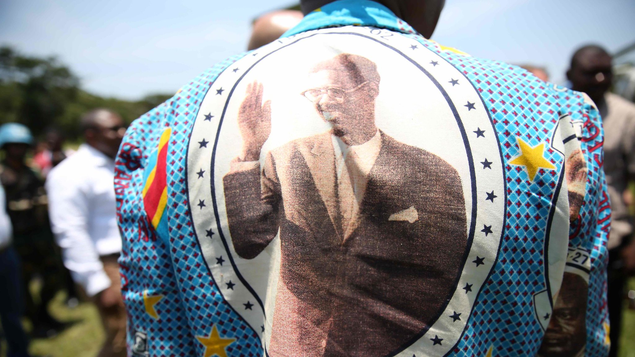 Cette image montre une personne portant un grand vêtement aux motifs colorés et vifs. Le dos du vêtement est orné d'une grande impression d'une personne en costume, levant la main dans un geste de salut. Autour de l'image, on peut voir des étoiles et des motifs géométriques. La date "1961-01-17" est inscrite, indiquant probablement une signification importante liée à l'image. En arrière-plan, on aperçoit un groupe de personnes, suggérant un événement ou une réunion en extérieur sous un ciel bleu.