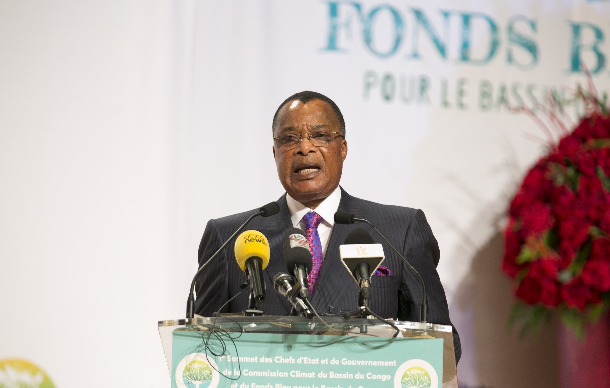 L'image montre un homme, probablement un leader ou un représentant politique, s'exprimant devant un public. Il porte un costume sombre avec une cravate rose et semble être en train de donner un discours. Devant lui, il y a un podium avec plusieurs microphones et un panneau indiquant "Fonds Bleu pour le Bassin du Congo". En arrière-plan, on peut voir des éléments décoratifs qui suggèrent un événement officiel, avec des logos et des affiches liés au thème de la conservation et du développement. L'atmosphère semble sérieuse, indiquant l'importance de l'événement.
