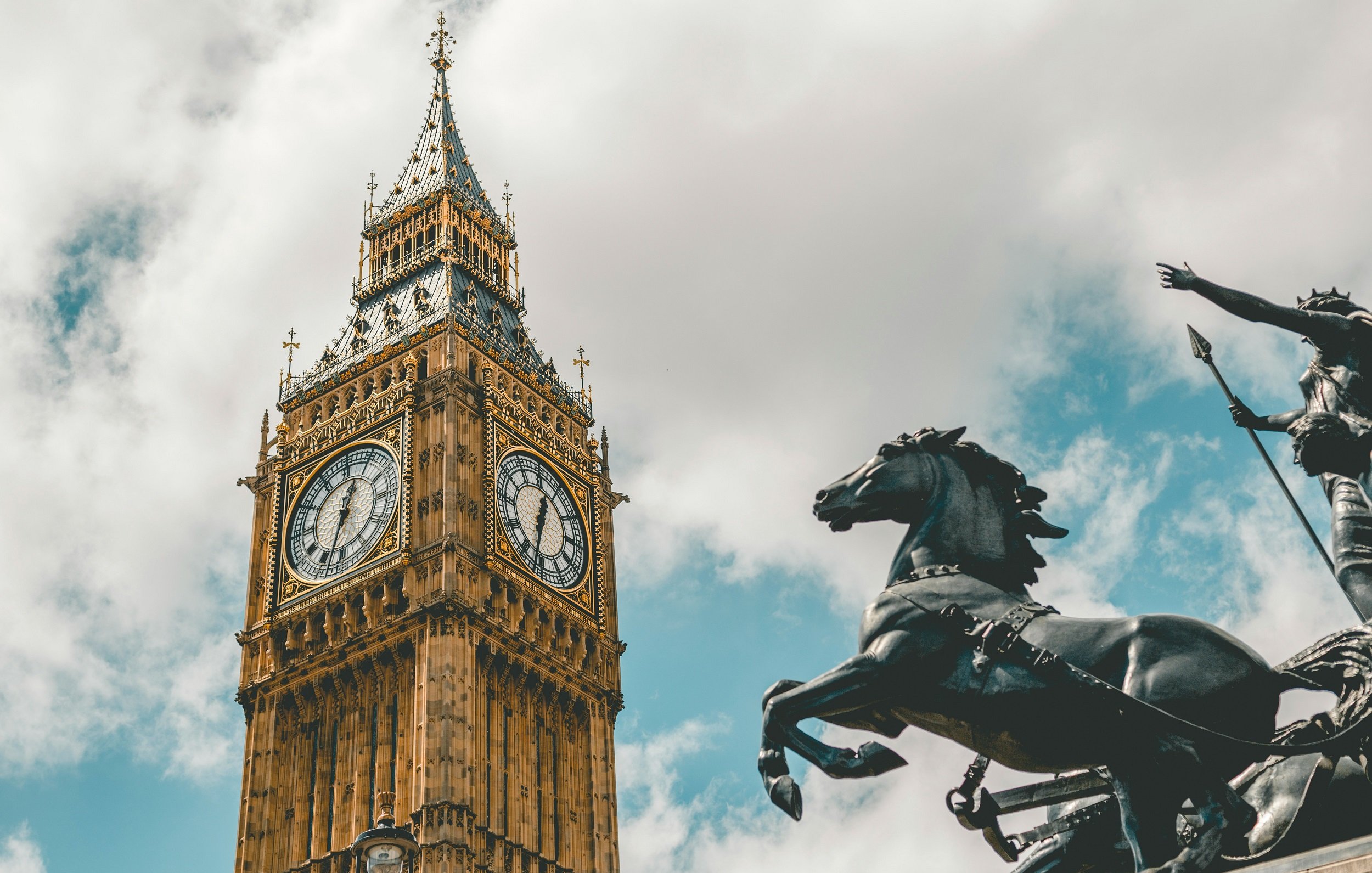 L'image présente une vue majestueuse de la célèbre horloge de Big Ben à Londres. Le bâtiment, en pierre, se distingue par sa hauteur impressionnante et ses ornements dorés. Les deux grands cadrans d'horloge sont visibles, indiquant l'heure avec des chiffres romains. En bas à droite, une statue en bronze d'un cavalier sur un cheval ajoute une touche historique et dynamique à la scène. Le ciel est partiellement nuageux, ce qui crée un contraste intéressant avec l'architecture dorée et les détails du monument. L'ensemble dégage une atmosphère à la fois emblématique et intemporelle.