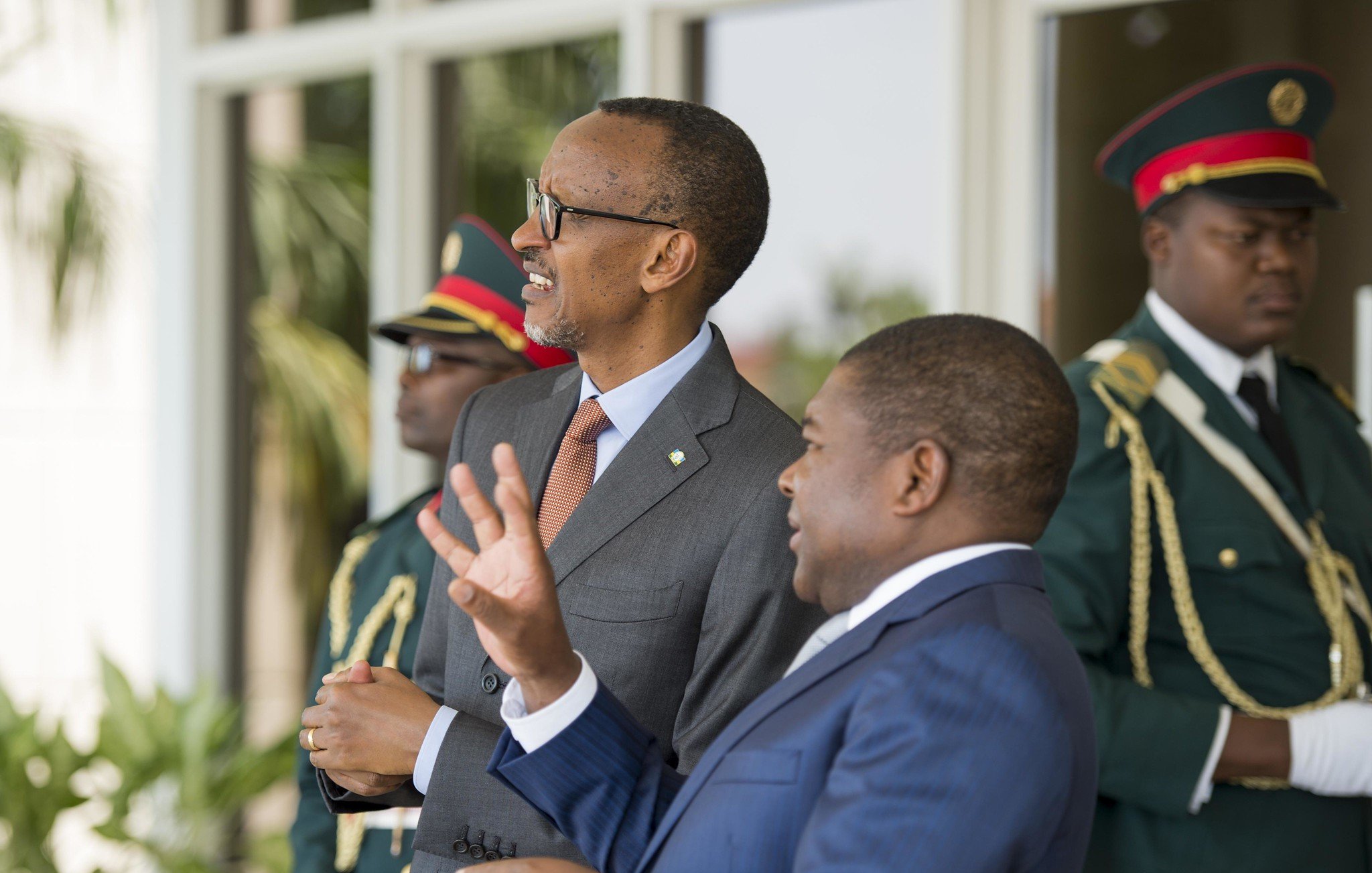 Sur cette image, nous voyons deux hommes en costume dialoguant dans un cadre officiel. L'homme à gauche, avec des lunettes et un sourire, semble être en train d'écouter attentivement. À sa droite, un homme aux cheveux courts gesticule avec les mains, soulignant un point dans la conversation. En arrière-plan, deux gardes en uniforme militaire se tiennent, assurant une ambiance de sécurité. On peut également percevoir des éléments de verdure, suggérant un environnement extérieur agréable.
