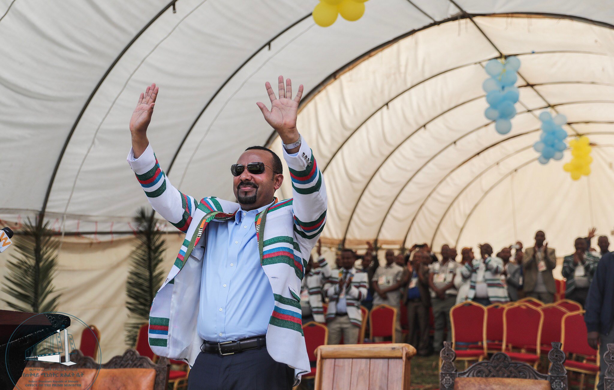 L'image montre un homme, probablement un leader, se tenant debout sous un grand chapiteau. Il porte une tenue colorée avec des rayures multicolores et des lunettes de soleil, et lève les deux mains dans un geste de salutation ou d'encouragement. En arrière-plan, on aperçoit un groupe de personnes qui semblent l'écouter attentivement, tous habillés de manière décontractée. Le chapiteau est décoré avec des ballons, ajoutant une ambiance festive à l'événement. L'atmosphère générale est celle d'un rassemblement joyeux et engageant.