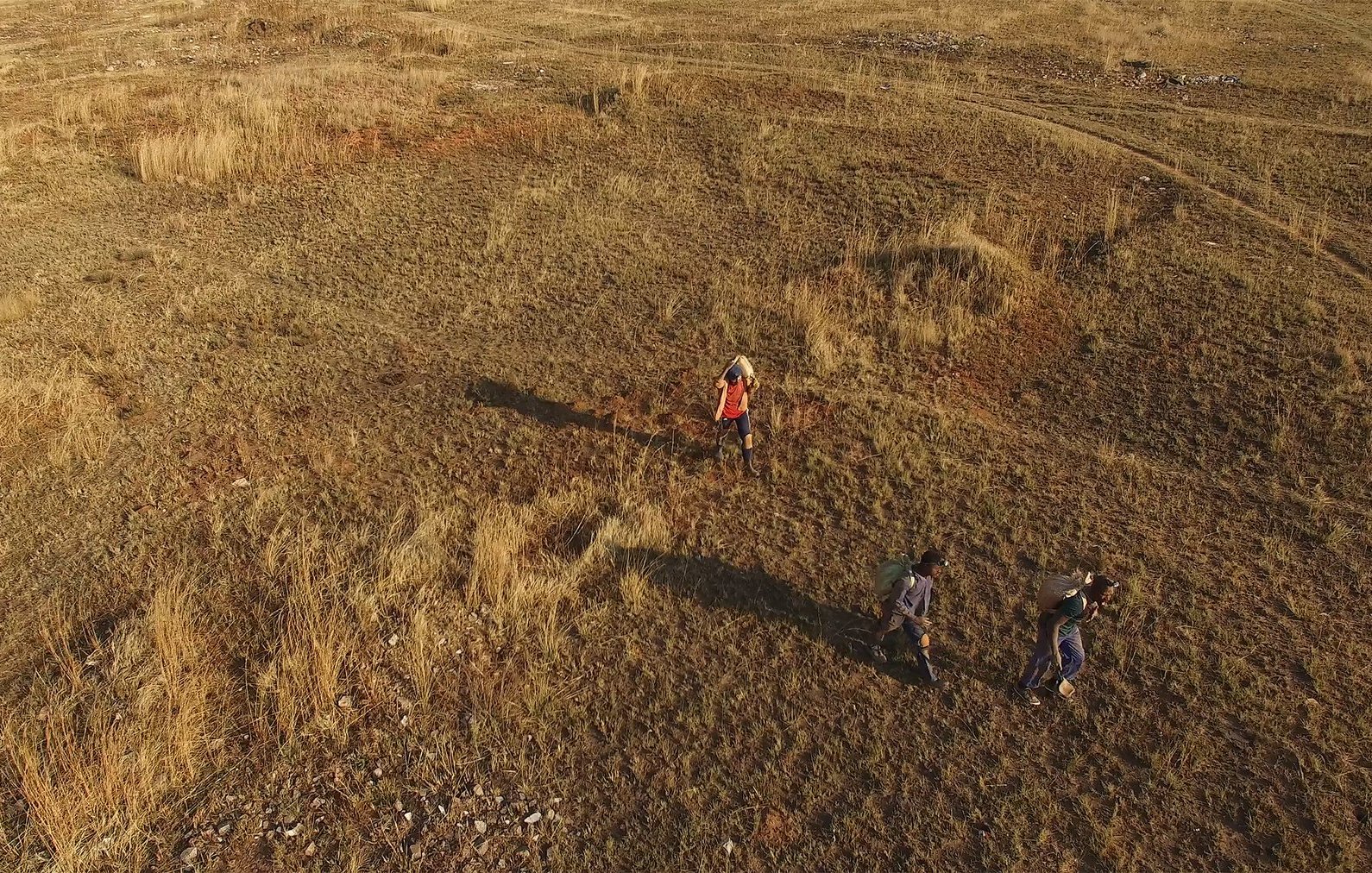 Dans cette image, on voit un paysage vaste et ouvert, couvert d'herbe sèche et de petites touffes de plantes. Le sol est légèrement accidenté, avec des zones de terre claire et quelques pierres éparpillées. Trois personnes marchent dans ce terrain. L'une d'elles porte un vêtement rouge, tandis que les autres sont habillées de couleurs plus sombres. Leur ombre s'étend sur le sol, suggérant que le soleil est assez bas dans le ciel. L'atmosphère est calme et naturelle, évoquant une promenade en pleine nature.