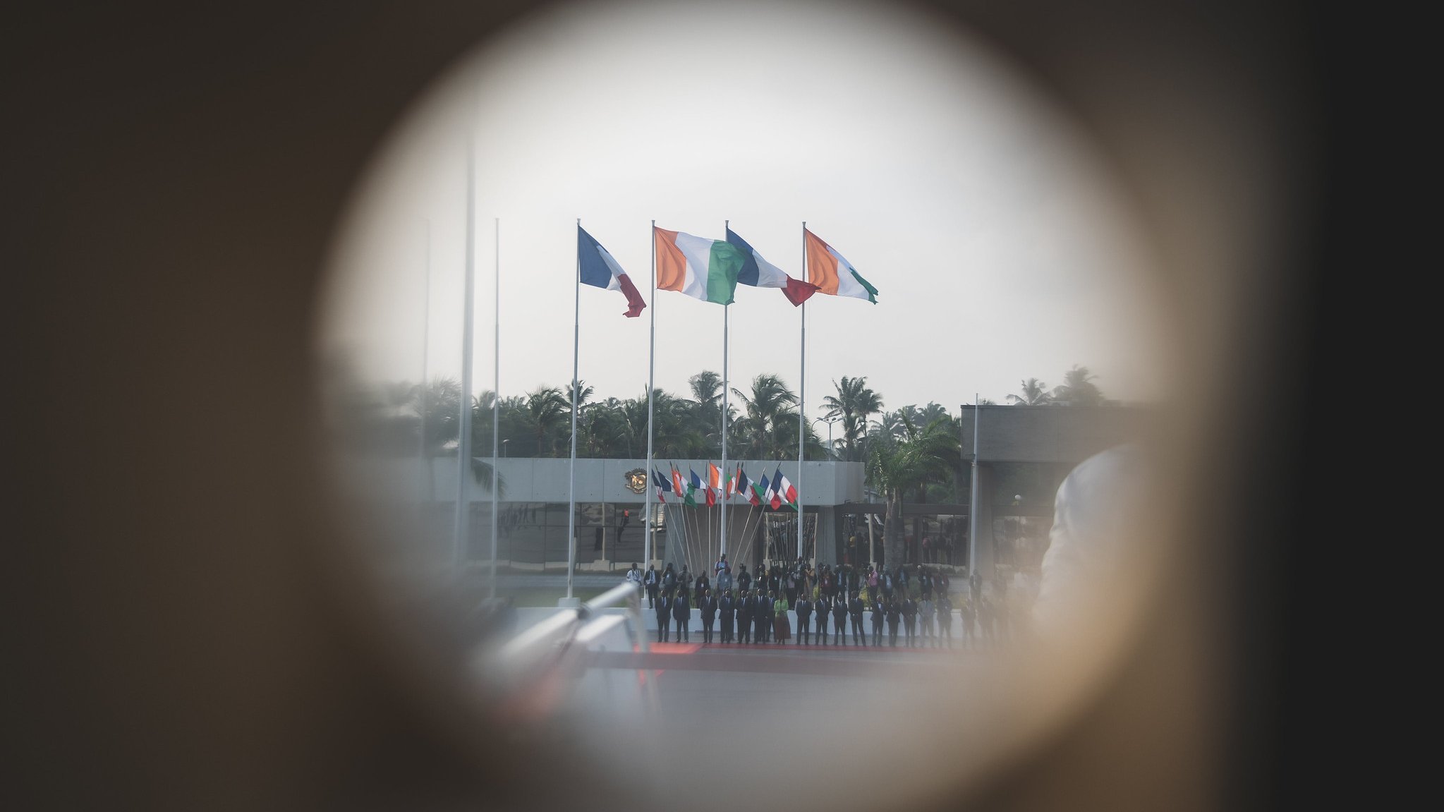 L'image montre un groupe de personnes alignées, probablement en uniforme, au centre d'un événement officiel. En arrière-plan, plusieurs drapeaux flottent au vent, représentant probablement différents pays. On distingue une ambiance solennelle, renforcée par la présence d'arbres aux feuilles verdoyantes et d'un espace dégagé. L'image est prise à travers un trou de serrure, ce qui donne une perspective unique et crée une atmosphère d'intimité et de mystère. La lumière semble douce et diffuse, suggérant un moment calme dans une journée ensoleillée.