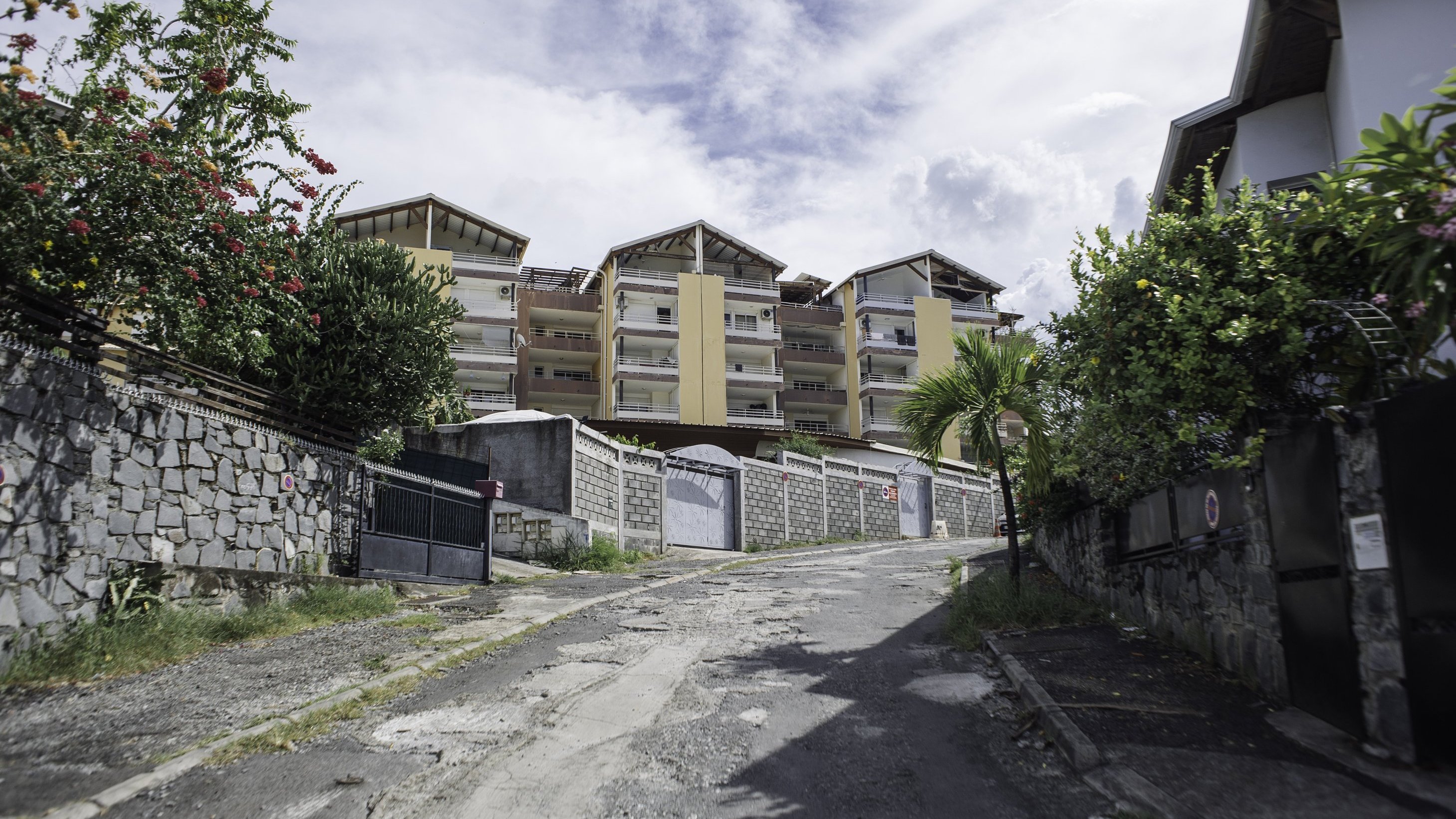 L'image montre une rue en pente, bordée de murs en pierre et de verdure. Sur le côté, on peut distinguer des fleurs colorées et des plantes tropicales. À l'arrière-plan, plusieurs bâtiments modernes avec des balcons se dressent, peints dans des teintes de jaune et de beige. Le ciel est nuageux, ce qui donne une ambiance légèrement dramatique à la scène. Le sol de la rue est inégal et montre des signes d'usure, avec des pavés qui apportent une texture variée au passage. L'atmosphère est calme, typique d'un quartier résidentiel.