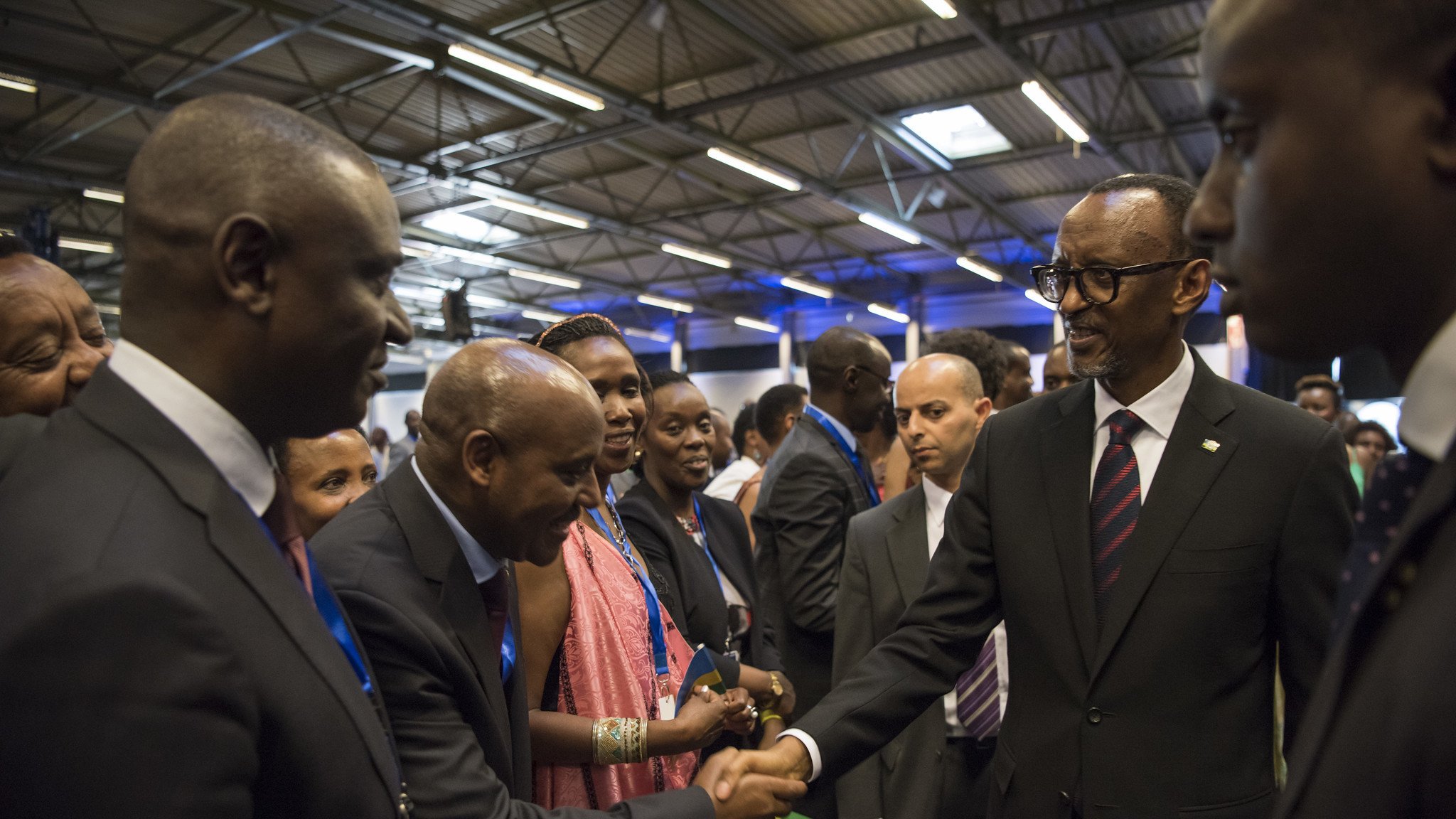 Dans cette image, nous voyons un groupe de personnes rassemblées dans un espace intérieur, probablement lors d'un événement officiel ou d'une conférence. Parmi eux se trouve un homme en costume noir avec une cravate, qui semble être un dirigeant ou une personne importante. Il tend la main pour saluer un autre homme, également en costume. Les autres personnes présentes, principalement habillées formellement, regardent avec intérêt. L'atmosphère est conviviale et respectueuse, avec des sourires et des échanges amicaux. Les murs sont décorés de manière simple, et un éclairage doux illumine la scène.