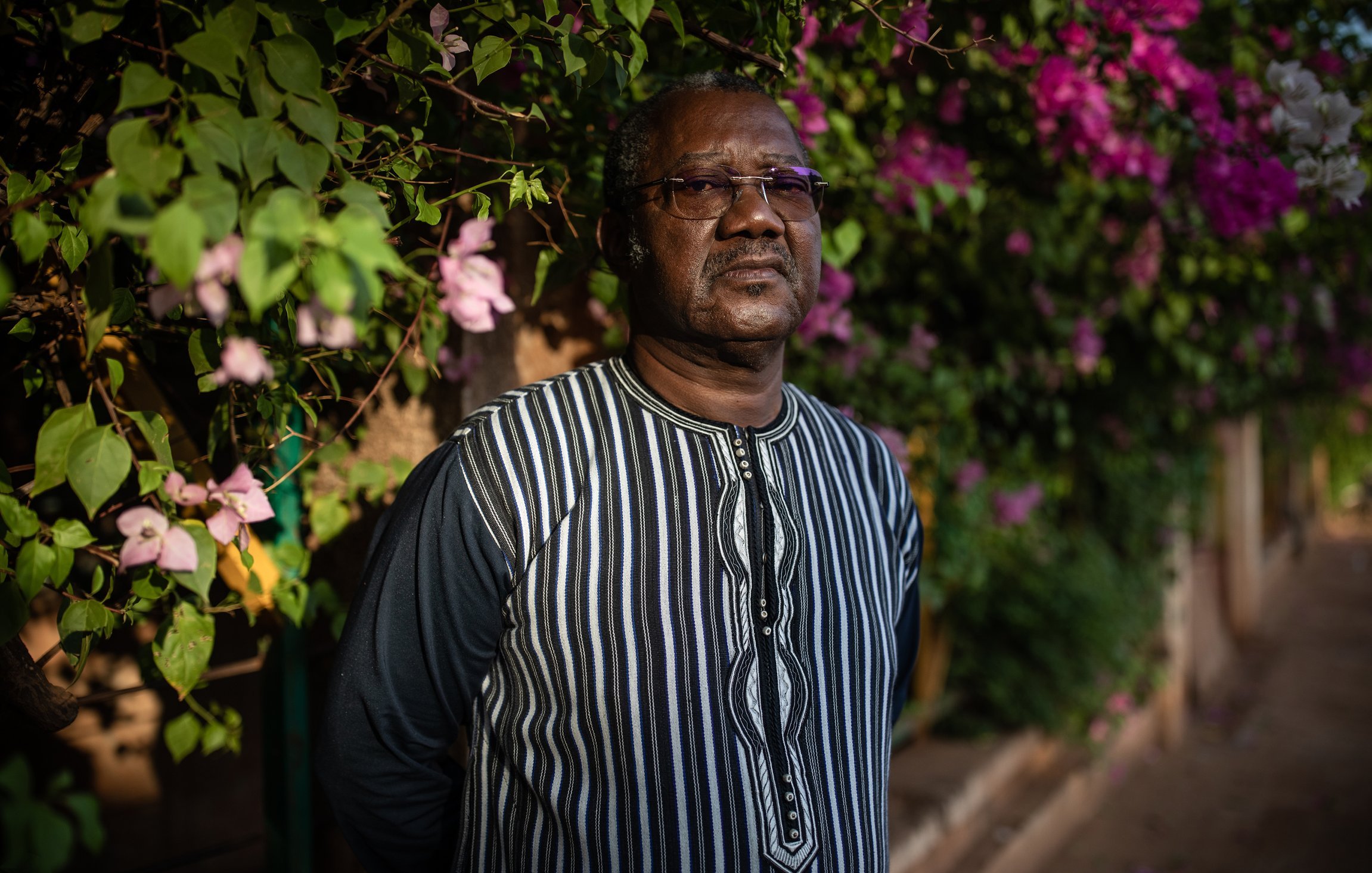 L'image montre un homme debout devant un mur recouvert de magnifiques bougainvilliers aux couleurs vives, principalement en rose et violet. Il porte une chemise à rayures noires et blanches, avec un col qui attire l'attention. Son expression est sérieuse et réfléchie, et il semble absorbé par ses pensées. En arrière-plan, on peut percevoir une ambiance douce et chaleureuse grâce à la lumière naturelle qui illumine la scène, accentuant la beauté des fleurs.