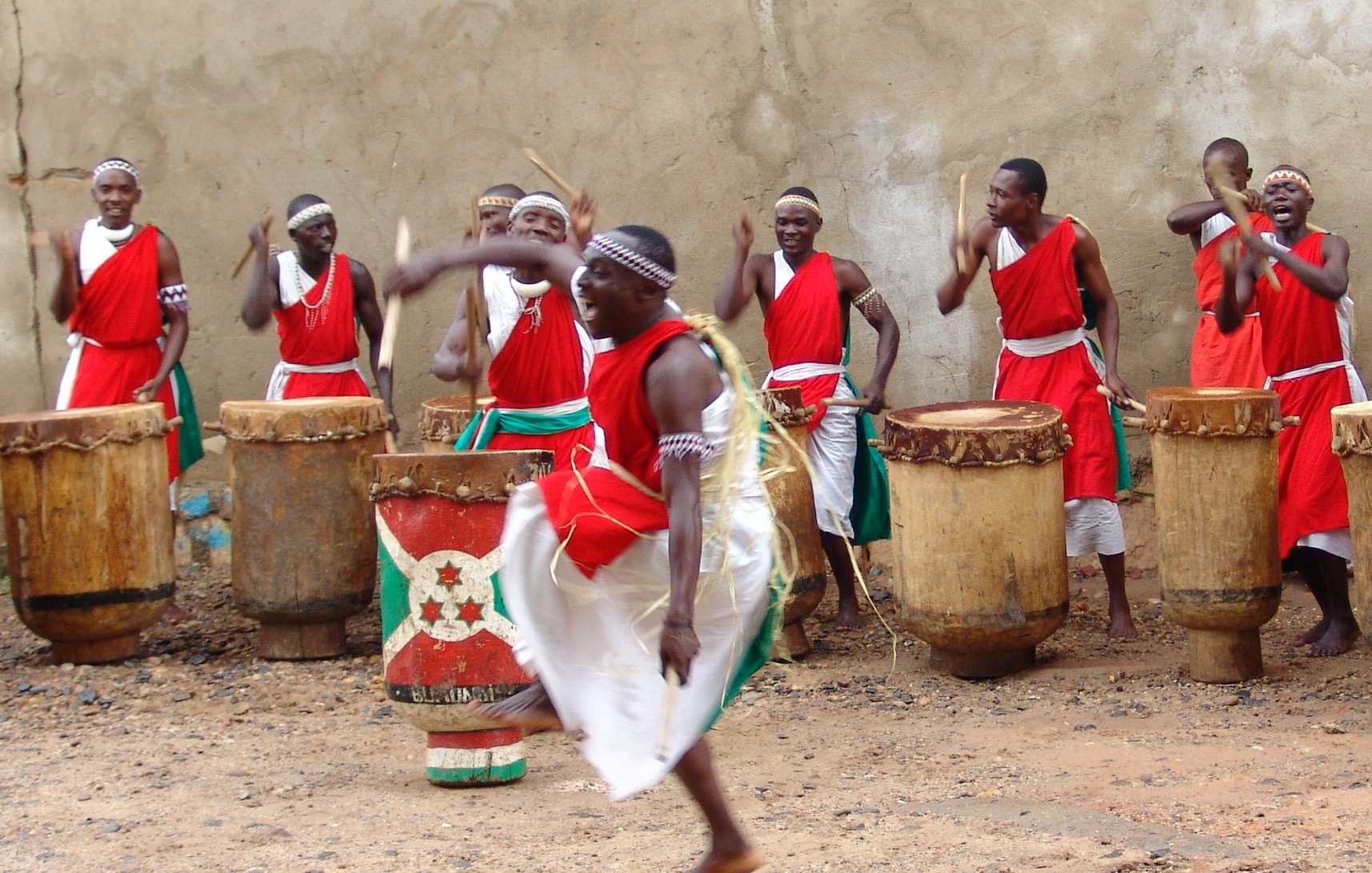 L'image représente un groupe de percussionnistes en pleine performance. Ils portent des vêtements traditionnels, principalement rouges, avec des ceintures vertes. Au centre, un homme se distingue en sautant avec énergie, créant un mouvement dynamique. Les autres membres du groupe, en arrière-plan, jouent des tambours en bois de forme cylindrique. L'atmosphère est festive et rythmée, avec une ambiance de célébration culturelle, où les sons des tambours résonnent fortement. Le sol est en terre, ce qui ajoute un aspect rustique à la scène.