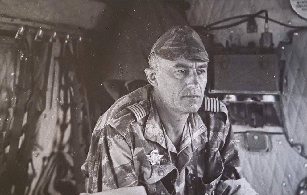 L'image présente un homme en uniforme militaire, assis à l'intérieur d'un véhicule. Il a les cheveux courts et grisonnants, avec une expression sérieuse et réfléchie. Son uniforme, camouflé, est orné d'insignes distinctifs sur les épaules, indiquant son grade. Au fond, on aperçoit des équipements militaires et des matériaux qui suggèrent un environnement de travail opérationnel. La lumière semble douce, et l'atmosphère générale dégage un sentiment de concentration et de détermination.