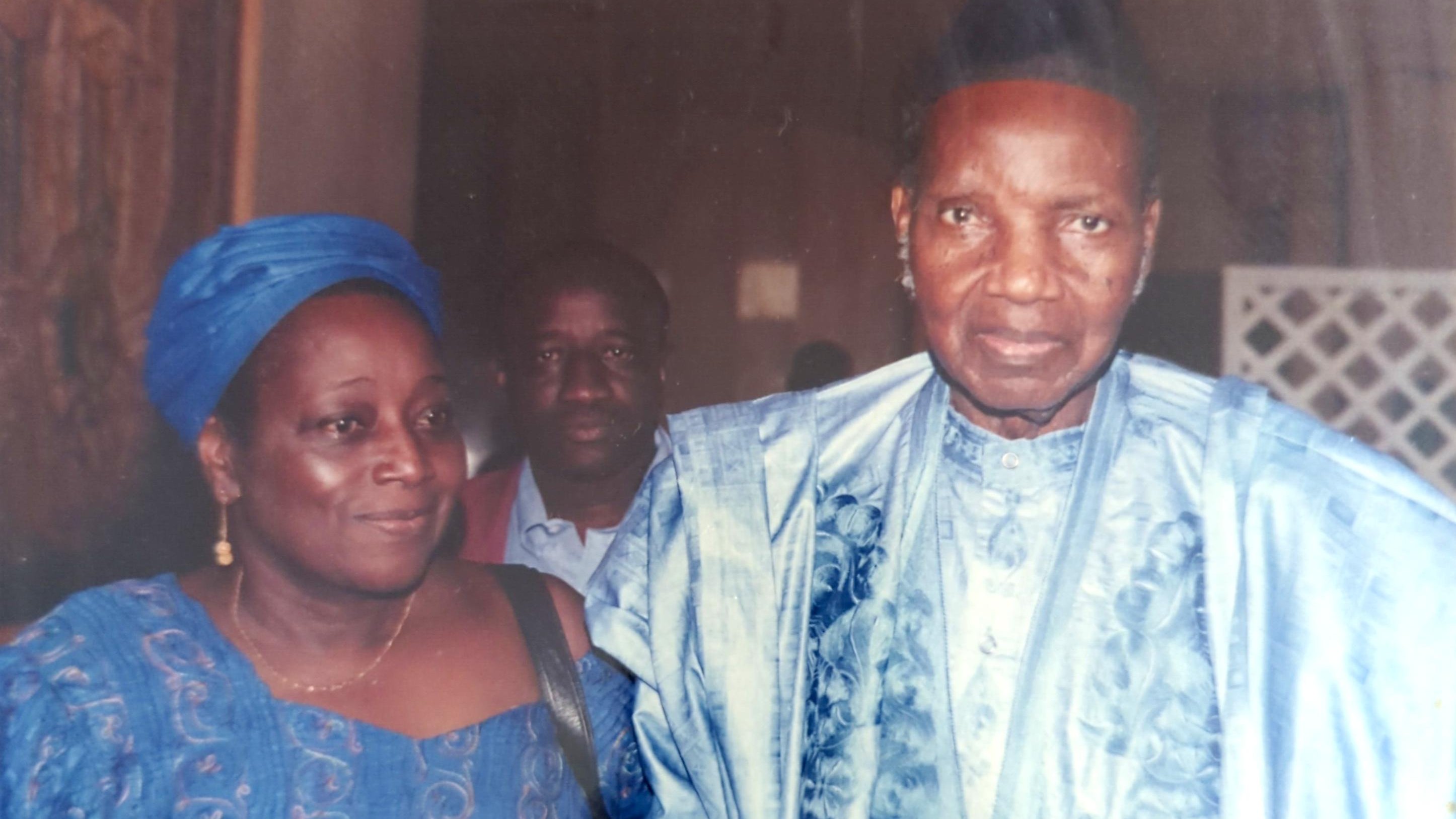 L'image montre trois personnes. À l'avant, un homme et une femme sont vêtus de costumes traditionnels, probablement en tissus d'importance culturelle. L'homme porte un chapeau et a un regard sérieux, tandis que la femme a un sourire et semble détendue. En arrière-plan, un troisième homme est visible, bien qu'il soit moins en avant-plan. L'ambiance de l'image semble formelle.