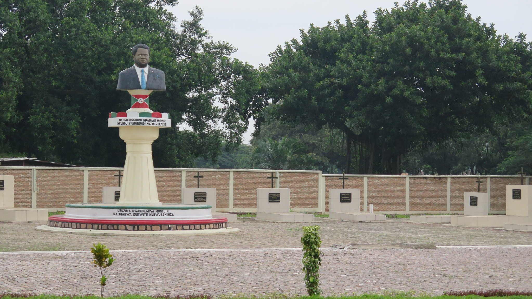 Cette image montre une statue en l'honneur d'une personnalité importante, probablement un leader ou un héros national. La statue est placée sur un socle élevé et représente un homme, vêtu d'un costume. En arrière-plan, on peut voir un mur avec plusieurs croix, qui semblent être des stèles commémoratives. Le site est entouré de verdure, avec des arbres touffus qui apportent une atmosphère paisible. Le sol est en pavé, et des plantes décoratives sont présentes, ajoutant une touche de nature à cet espace de mémoire.