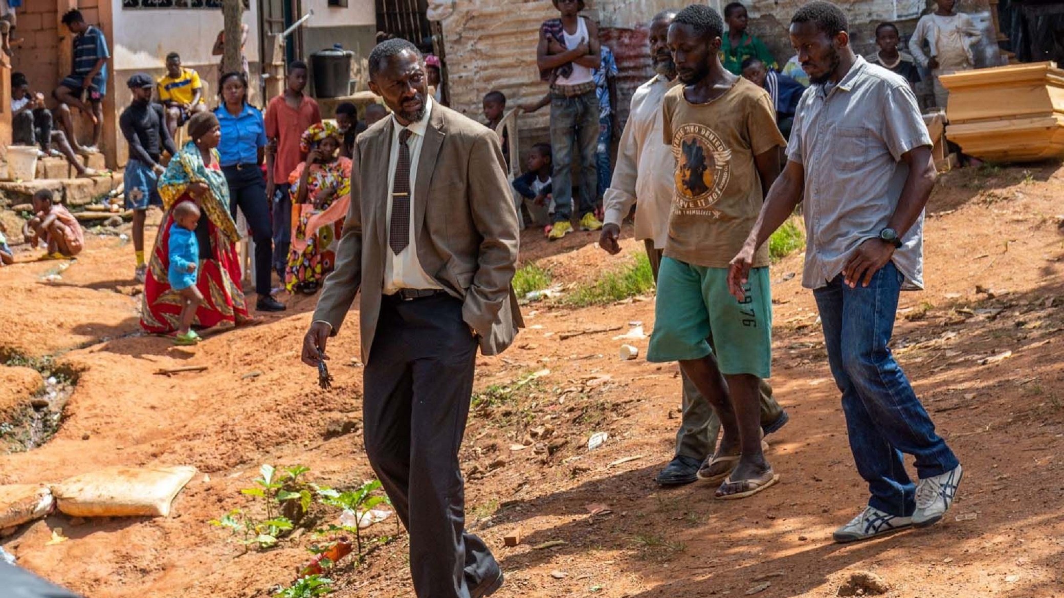 L'image montre un groupe de personnes dans un environnement rural ou semi-urbain. Au premier plan, un homme en costume cravate semble marcher avec assurance, le regard concentré. À sa gauche, un homme portant un t-shirt et un short se tient à côté de lui. D'autres personnes, habillées de manière décontractée, se déplacent également dans la scène, certains sont occupés à interagir entre eux. Le sol est en terre battue et il y a quelques plantes éparses. En arrière-plan, on peut apercevoir des maisons et des groupes de personnes qui discutent. L'atmosphère semble conviviale mais déterminée, avec un sentiment de communauté visible parmi les participants.