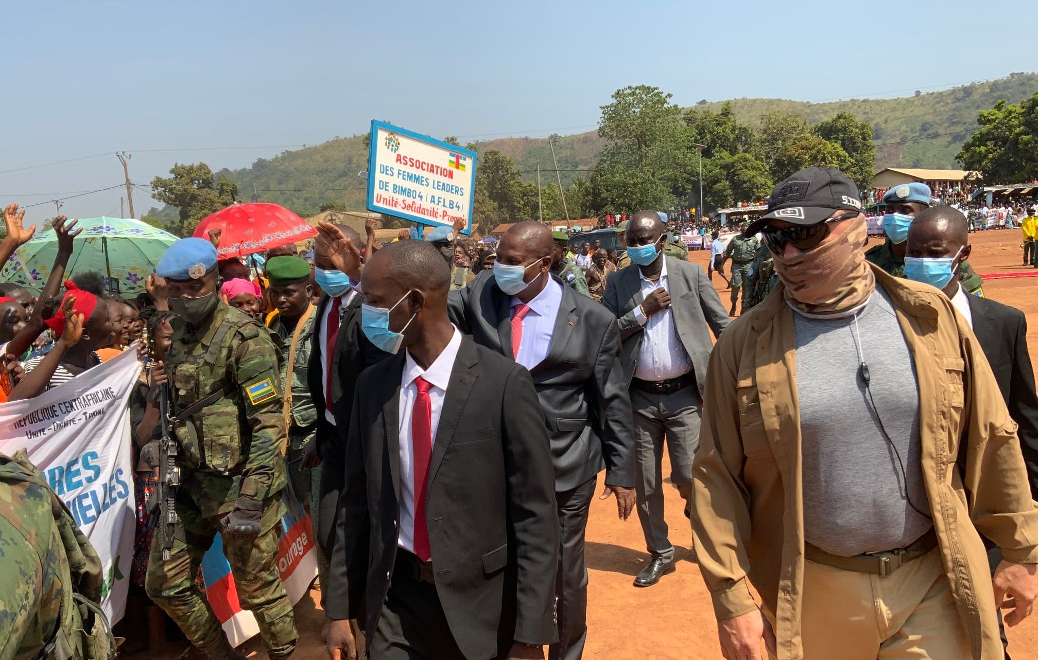 Dans cette image, un groupe de personnes est rassemblé lors d'un événement en extérieur. Au premier plan, on voit plusieurs hommes en costume, parmi lesquels un homme en particulier porte des lunettes de soleil et un foulard. Ils avancent au milieu d'une foule. À gauche, des militaires en uniforme surveillent la situation. En arrière-plan, des gens brandissent des pancartes, et des parasols colorés ajoutent une ambiance vivante à la scène. Le décor est verdoyant, avec des collines en arrière-plan sous un ciel clair. L'atmosphère semble festive et dynamique.