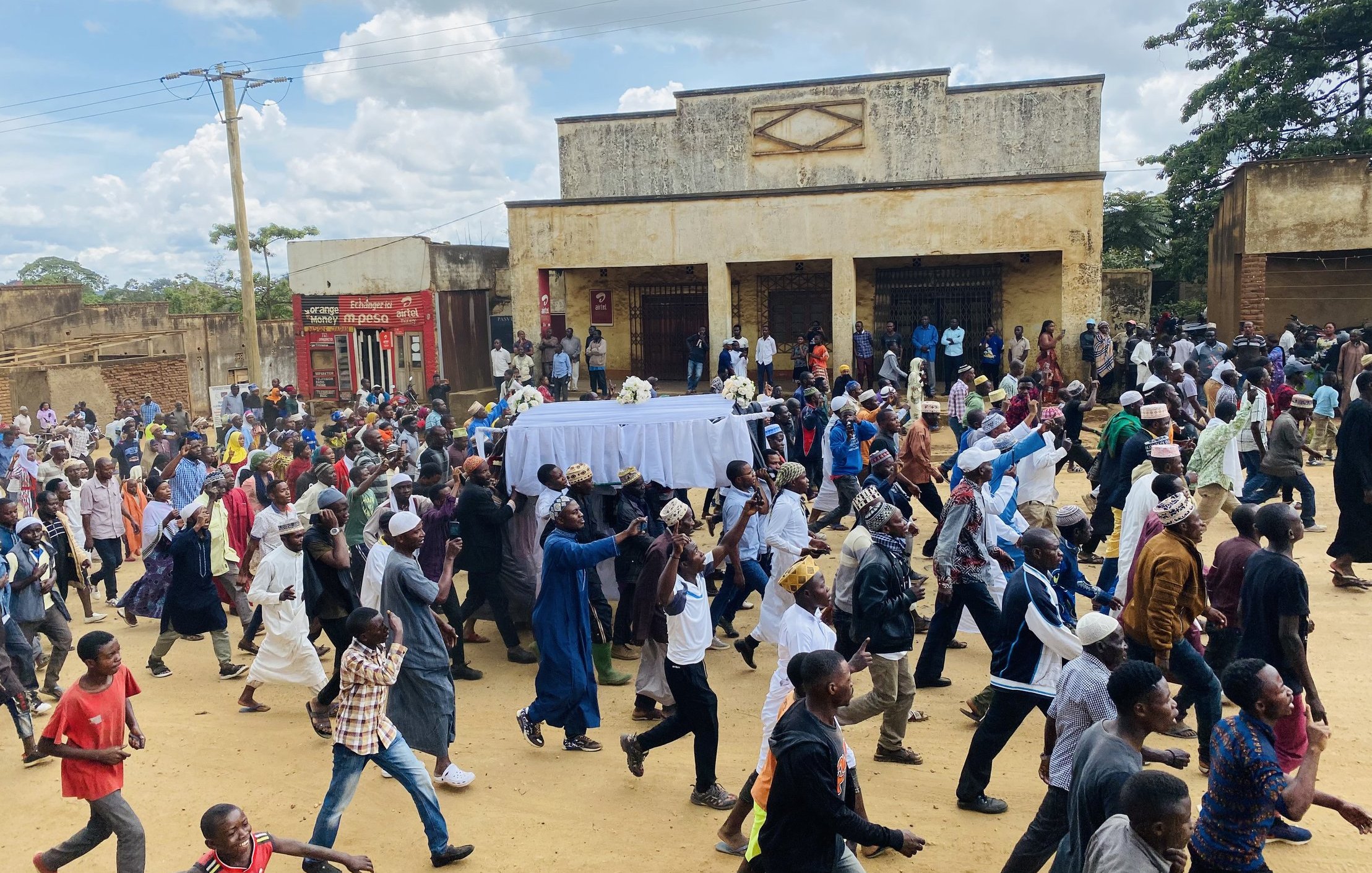 L'image montre une scène animée dans une rue où une grande foule se déplace. Au centre de l'attention, des personnes portent un drap blanc qui semble recouvrir un cercueil, indiquant qu'il s'agit d'un cortège funéraire. Les membres de la foule sont vêtus de divers habits, certains en tenues traditionnelles, d'autres plus contemporaines. Les visages reflètent des émotions variées, allant du recueillement à la tristesse. À l'arrière-plan, on peut remarquer des bâtiments aux murs fissurés, avec des panneaux publicitaires visibles. Le ciel est légèrement nuageux, créant une atmosphère à la fois sombre et lugubre, mais la scène reste vibrante de vie grâce à la présence de nombreux participants.