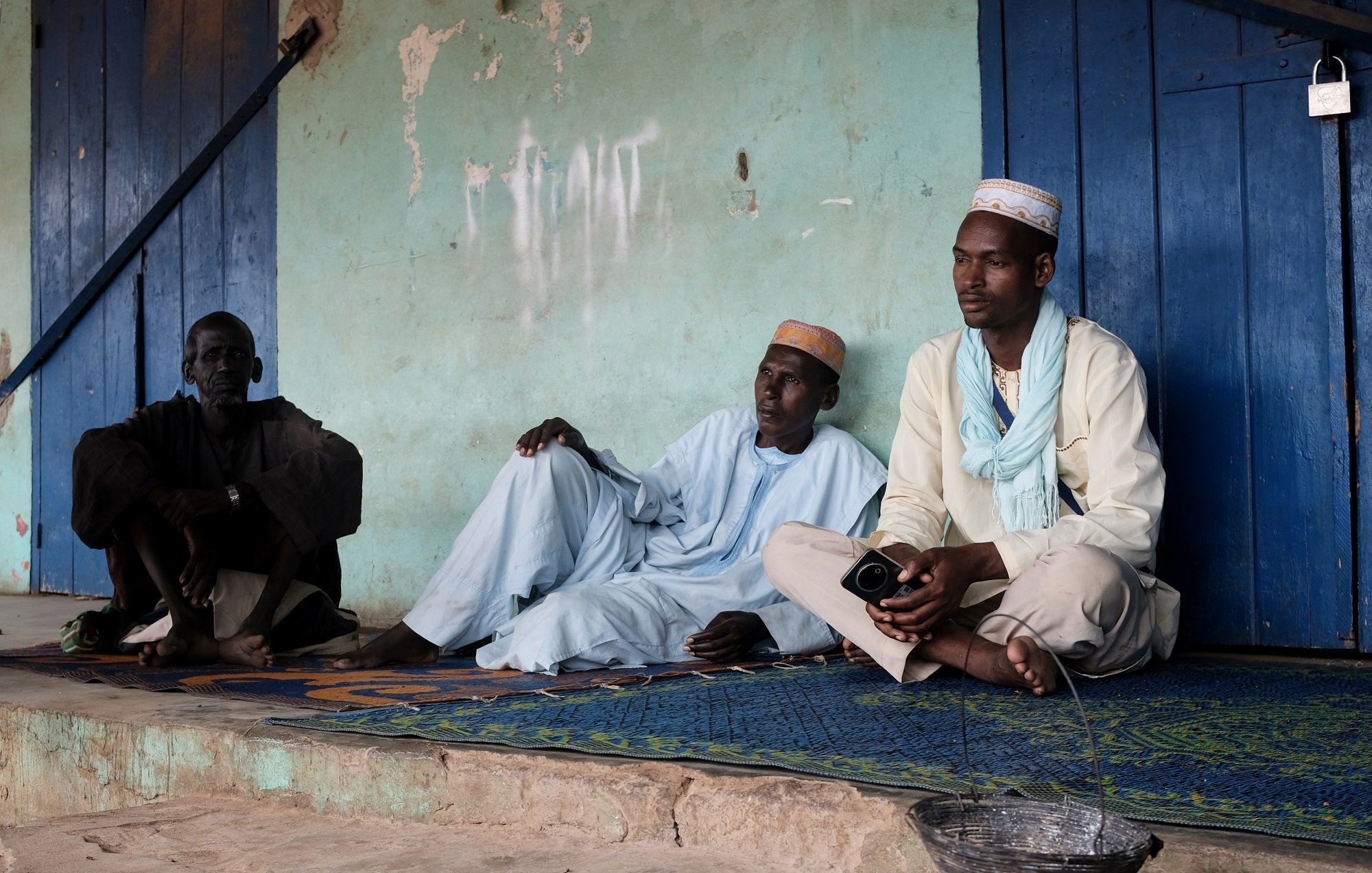 Cette image montre trois hommes assis sur un tapis, à l'extérieur d'un bâtiment aux murs de couleur turquoise. L'atmosphère semble calme et détendue. Ils portent des vêtements traditionnels : deux d'entre eux sont vêtus de tuniques, tandis que le troisième porte un chapeau. L'un des hommes est assis en tailleur, tandis que les deux autres sont en position plus détendue, le dos appuyé contre le mur. À côté d'eux, il y a un petit seau en métal d'où s'écoule un peu d'eau. En arrière-plan, des murs sont partiellement recouverts de graffiti. L'environnement semble rural, avec une lumière douce qui illumine la scène.