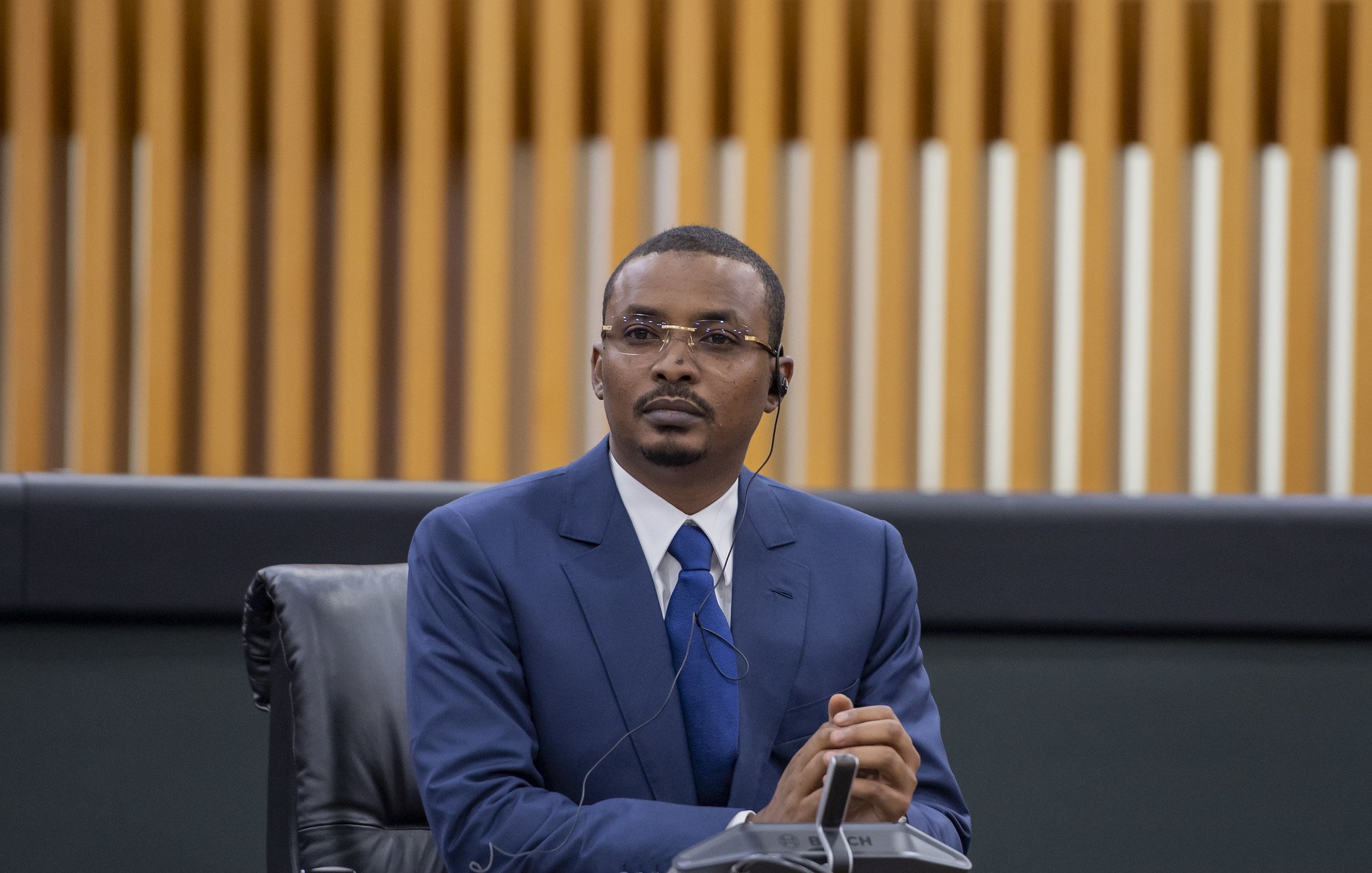 Dans cette image, un homme est assis à une table dans un environnement formel, probablement lors d'une réunion ou d'une conférence. Il porte un costume bleu, une chemise blanche et une cravate bleue. Ses cheveux sont courts et bien coiffés. L'homme semble concentré et attentif, avec un léger froncement de sourcils et les mains croisées devant lui. À son oreille, on peut voir un écouteur, ce qui suggère qu'il écoute probablement une traduction ou une intervention en cours. En arrière-plan, on distingue des panneaux en bois aux lignes verticales, ajoutant une touche moderne à l'espace. L'atmosphère générale est professionnelle et sérieuse.